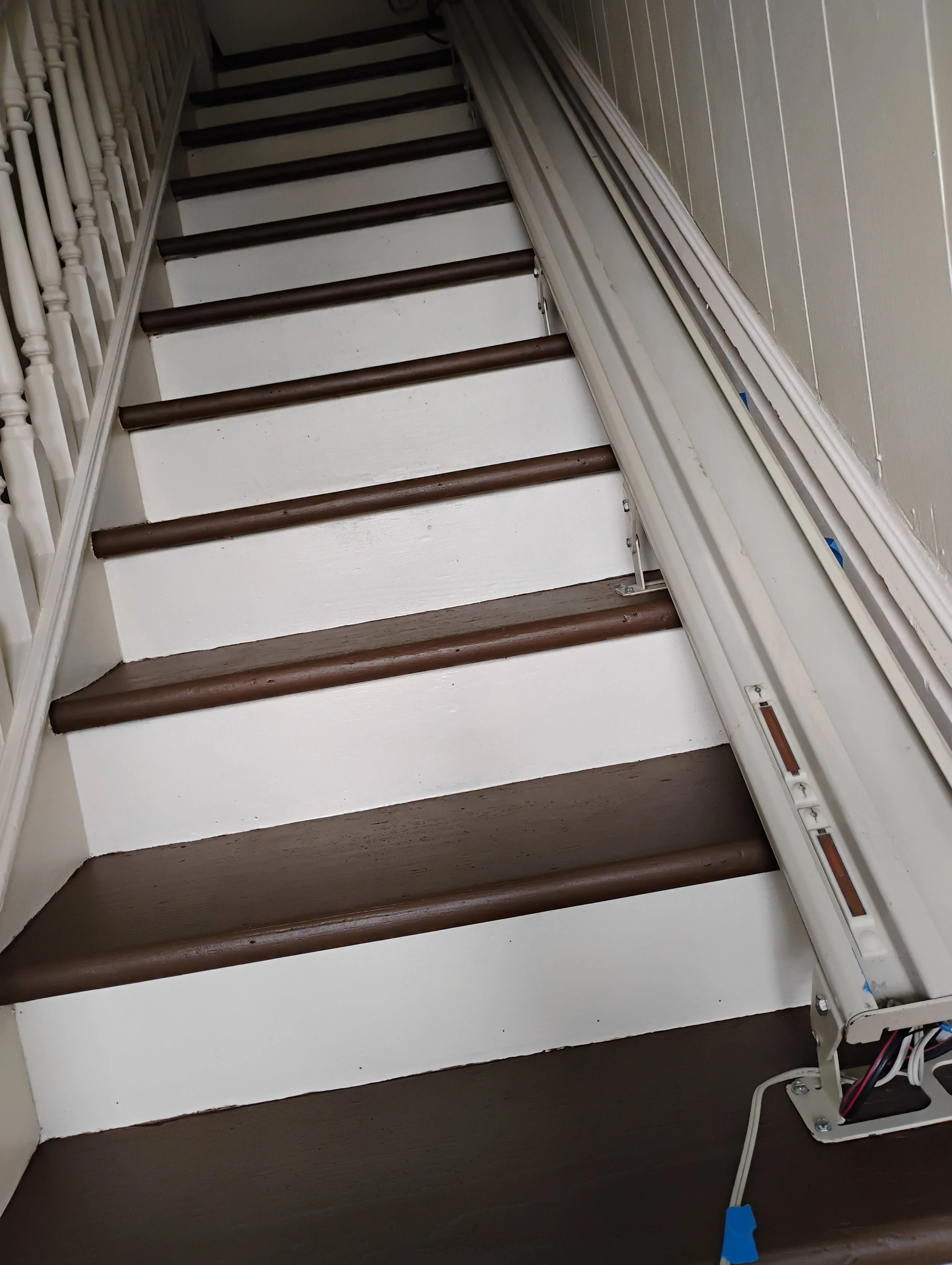 Staircase with white risers and dark brown treads, adjacent to a white wall and railing, with a heating or electrical unit on the side.