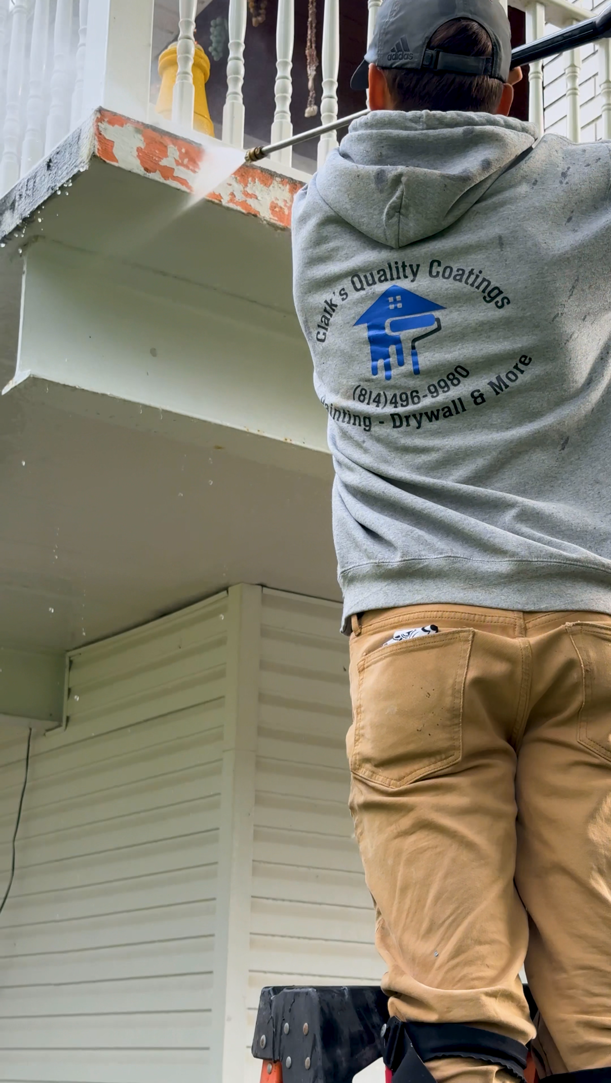 Worker pressure washing the underside of a porch or balcony with a gray hoodie that reads 'Clark's Quality Coatings' and a phone number, wearing khaki pants and a blue cap.