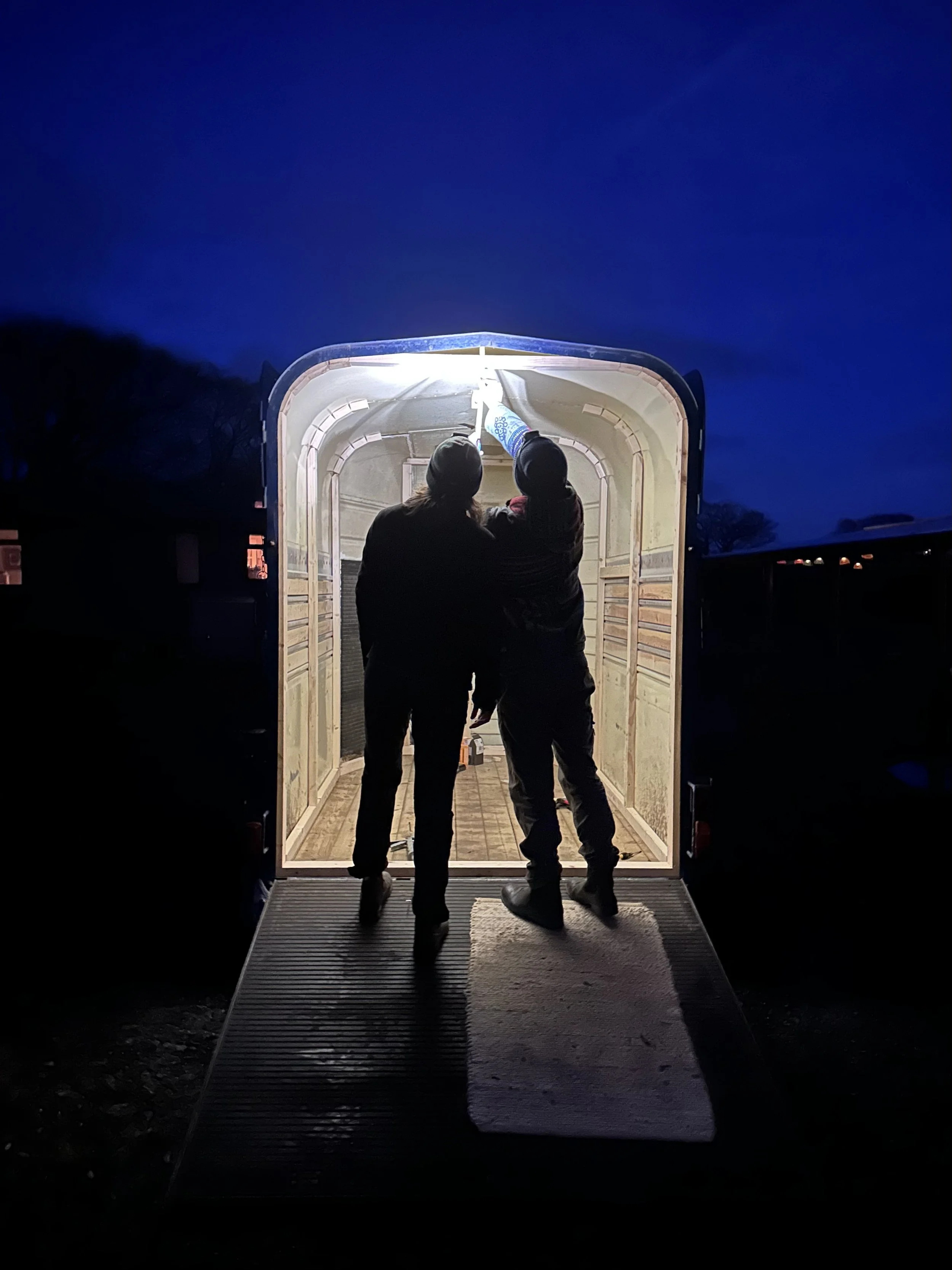 Cameron and Helena, silhouetted in the entrance of the sauna at night, durin the Sauna build but before anything was installed.  A dark blue sky in the background.