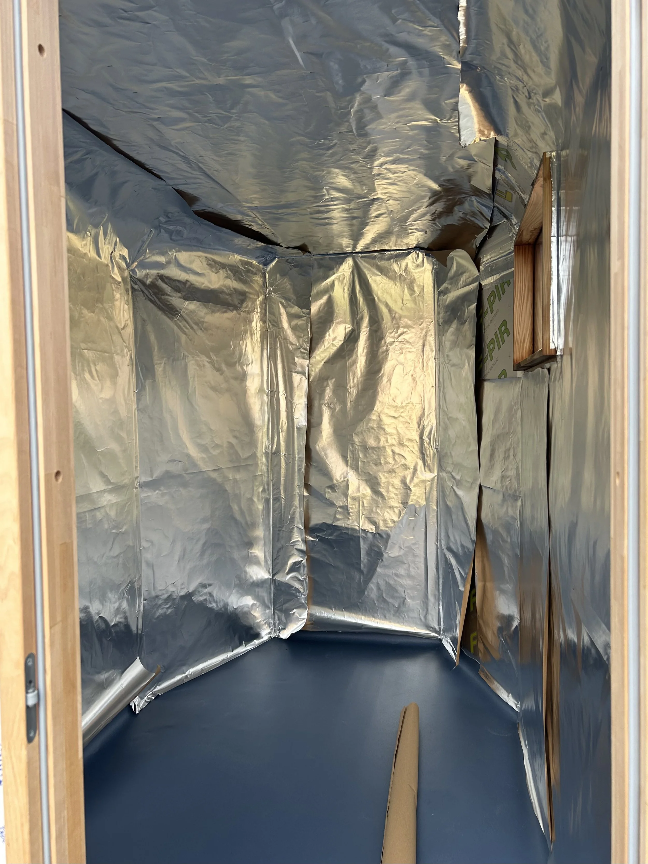 Empty interior of the drift sauna with reflective insulation covering the walls and ceiling and a wooden frame visible on the sides and window. pre cedar cladding, which is now installed at Livit adventure and glamping in Abbotsham, Bideford.
