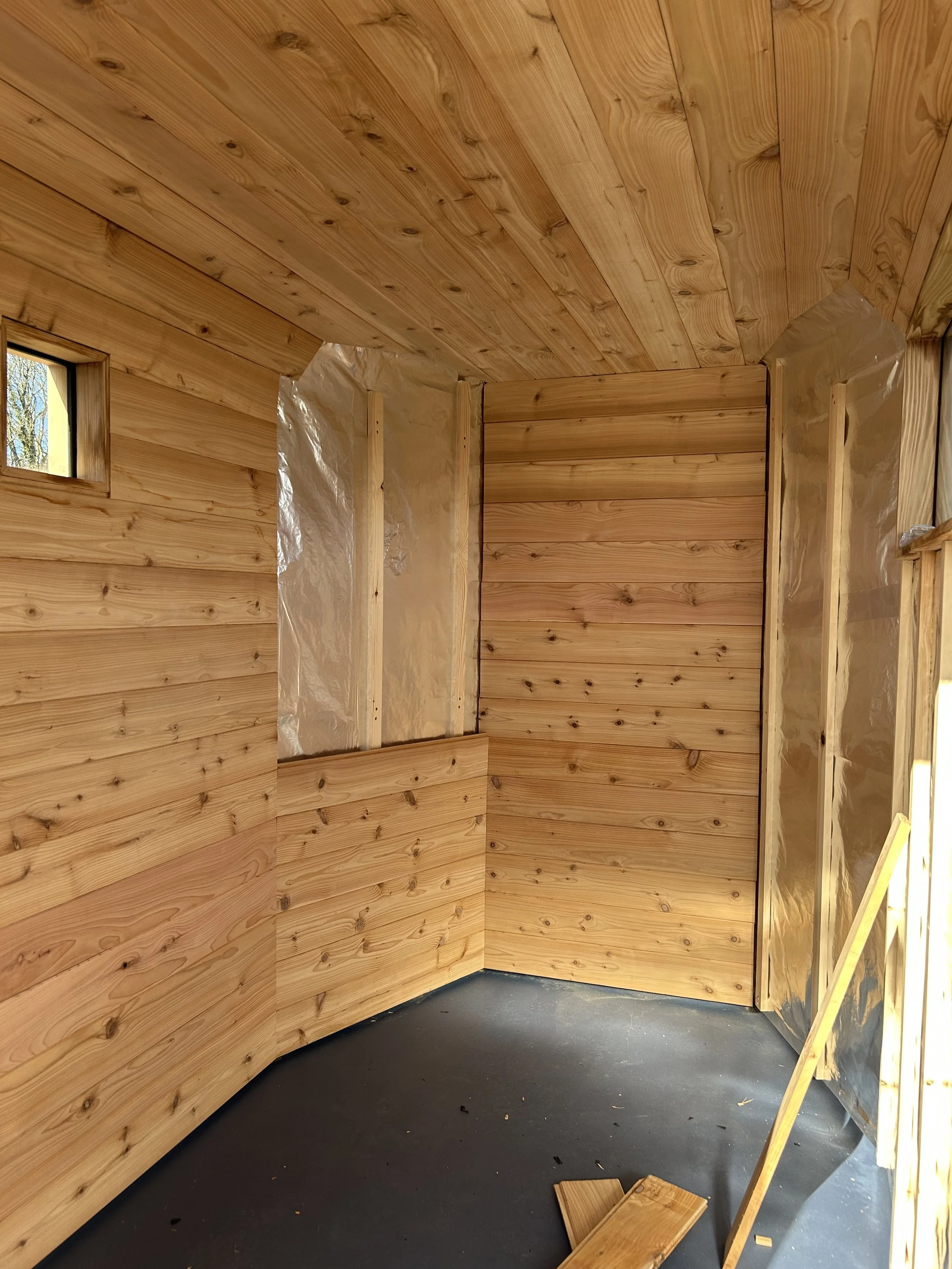 Interior of the Drift Sauna under construction with cedar cladding on walls and ceiling, a small window, and construction materials on the floor.