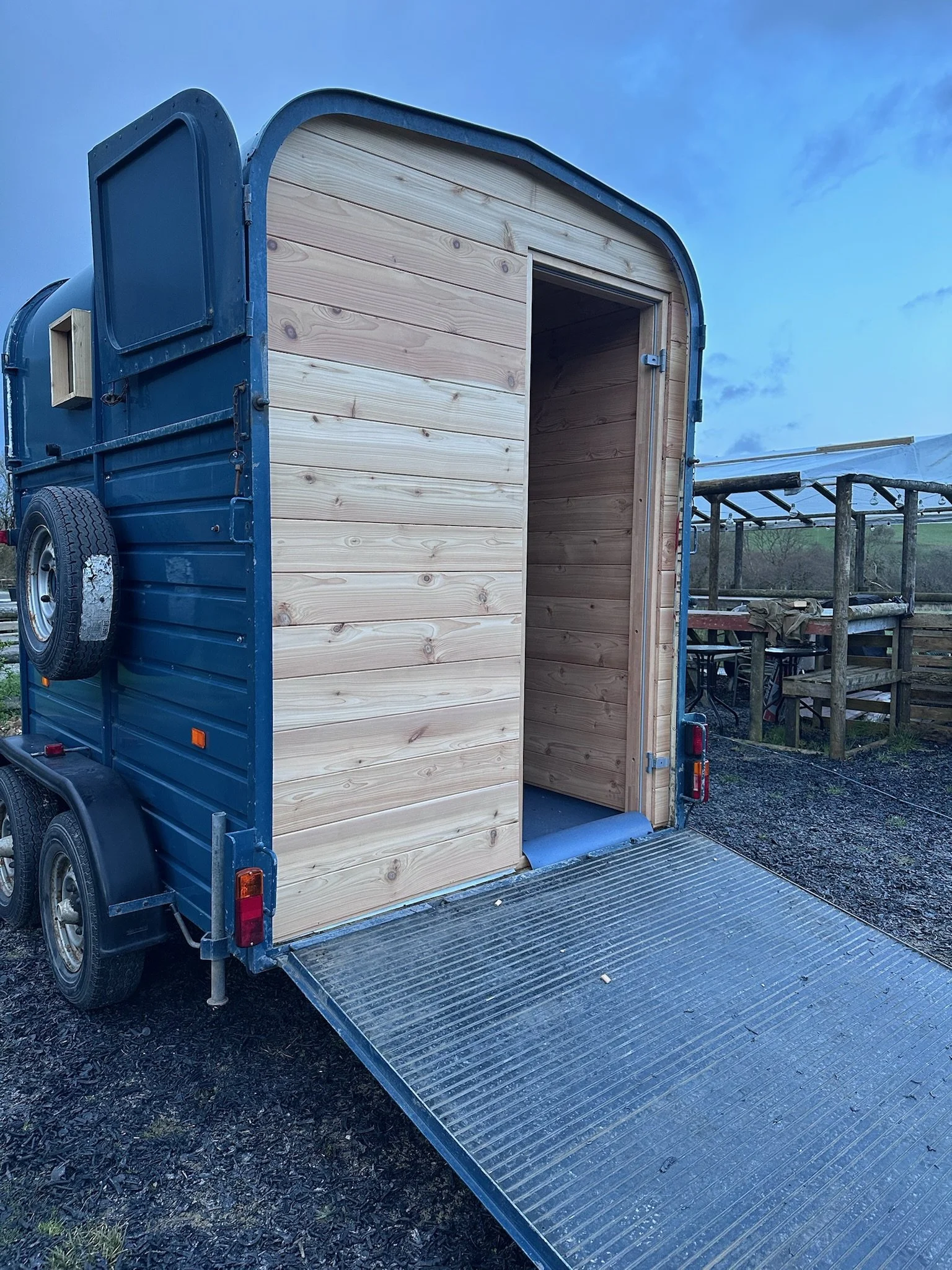 A small blue horse trailer with turned into a cedar sauna, the drift sauna. Image shows the ramp of the sauna at dusk, and the cedar cladding that lines it throughout.