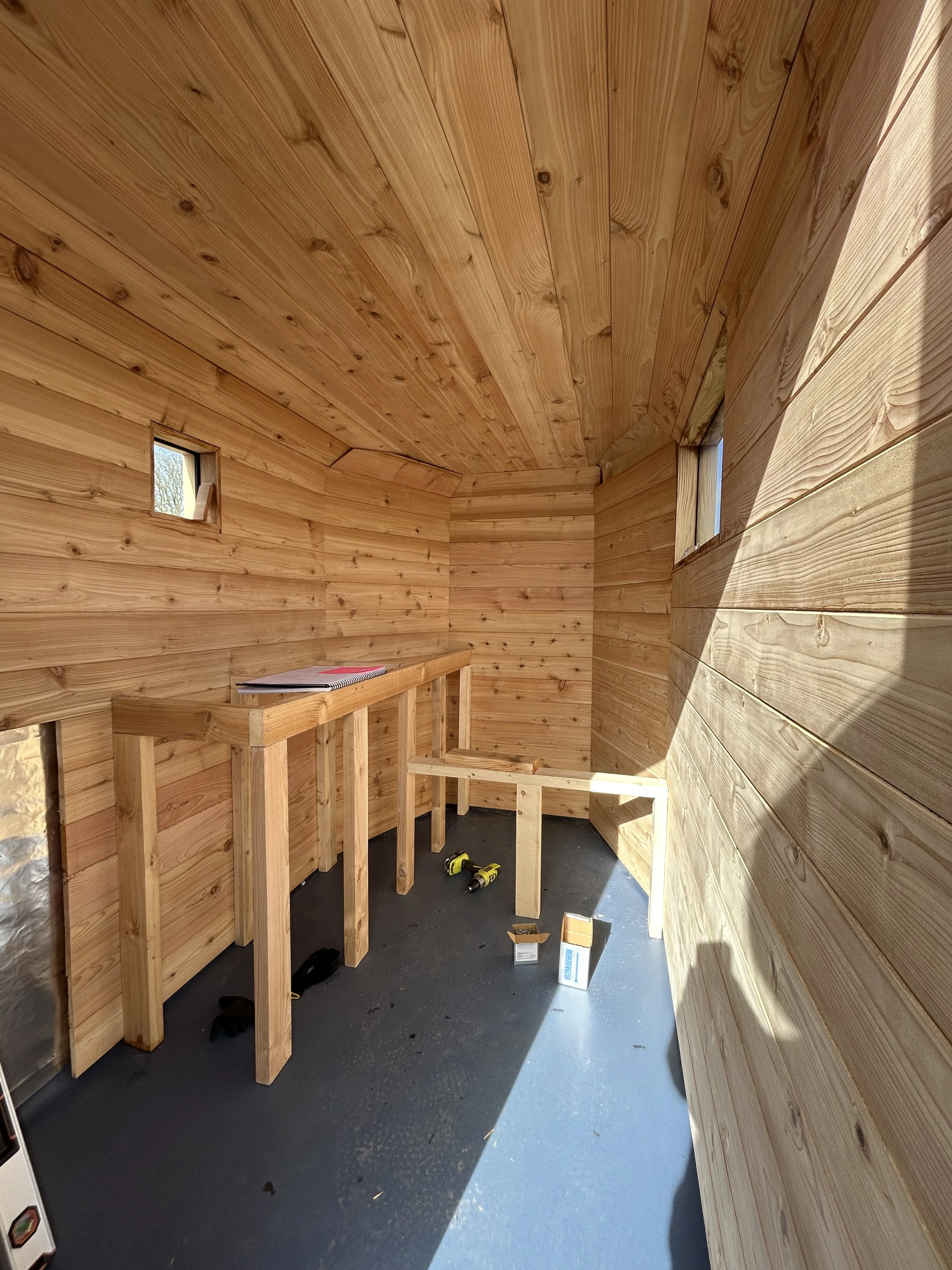 Cladded cedar walls and ceiling, small square windows, partially built cedar benches of the Drift Sauna, Bideford.