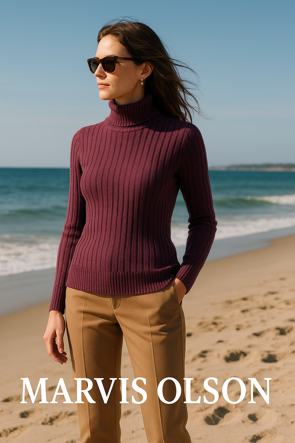 Woman in sunglasses wearing a maroon turtleneck sweater and tan pants standing on a beach by the ocean.