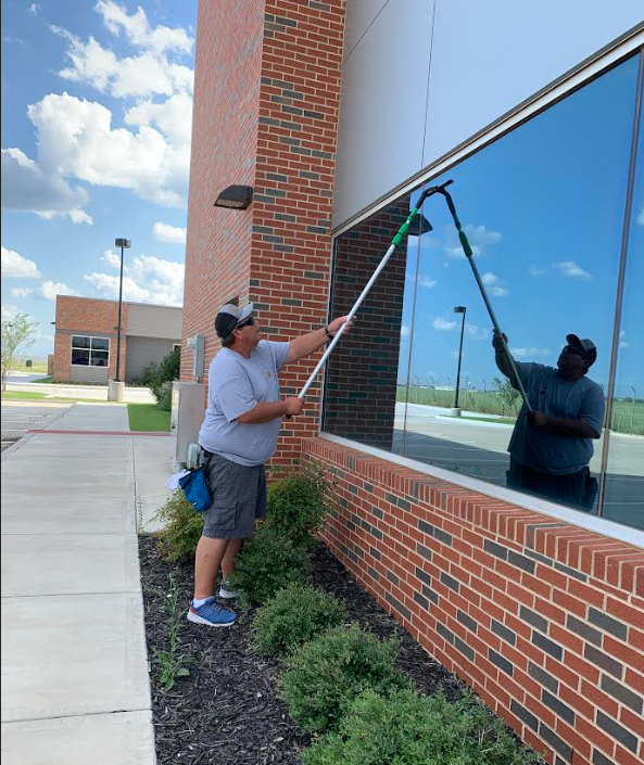 Man cleaning the outside glass windows of a commercial building with squeegees, standing on the sidewalk beside a brick wall with small bushes underneath.