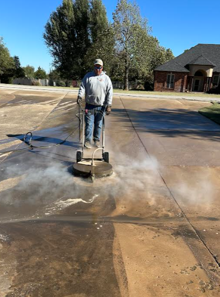 A man using pressure washing scrubber to clean driveway, in a residential neighborhood with trees and a house in the background.
