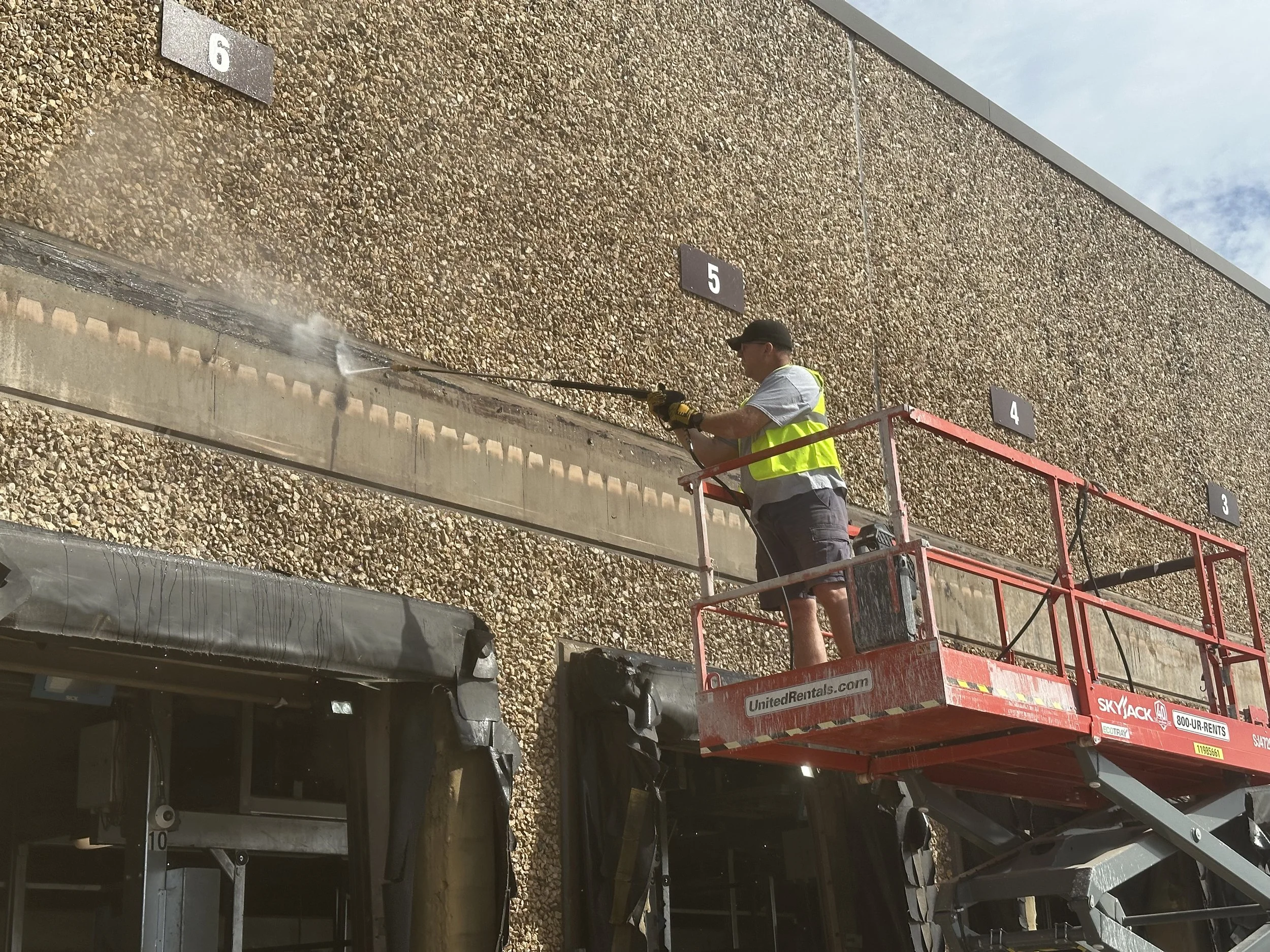 Worker in safety vest and hat cleaning the exterior wall of a building with a power washer from a cherry picker lift.