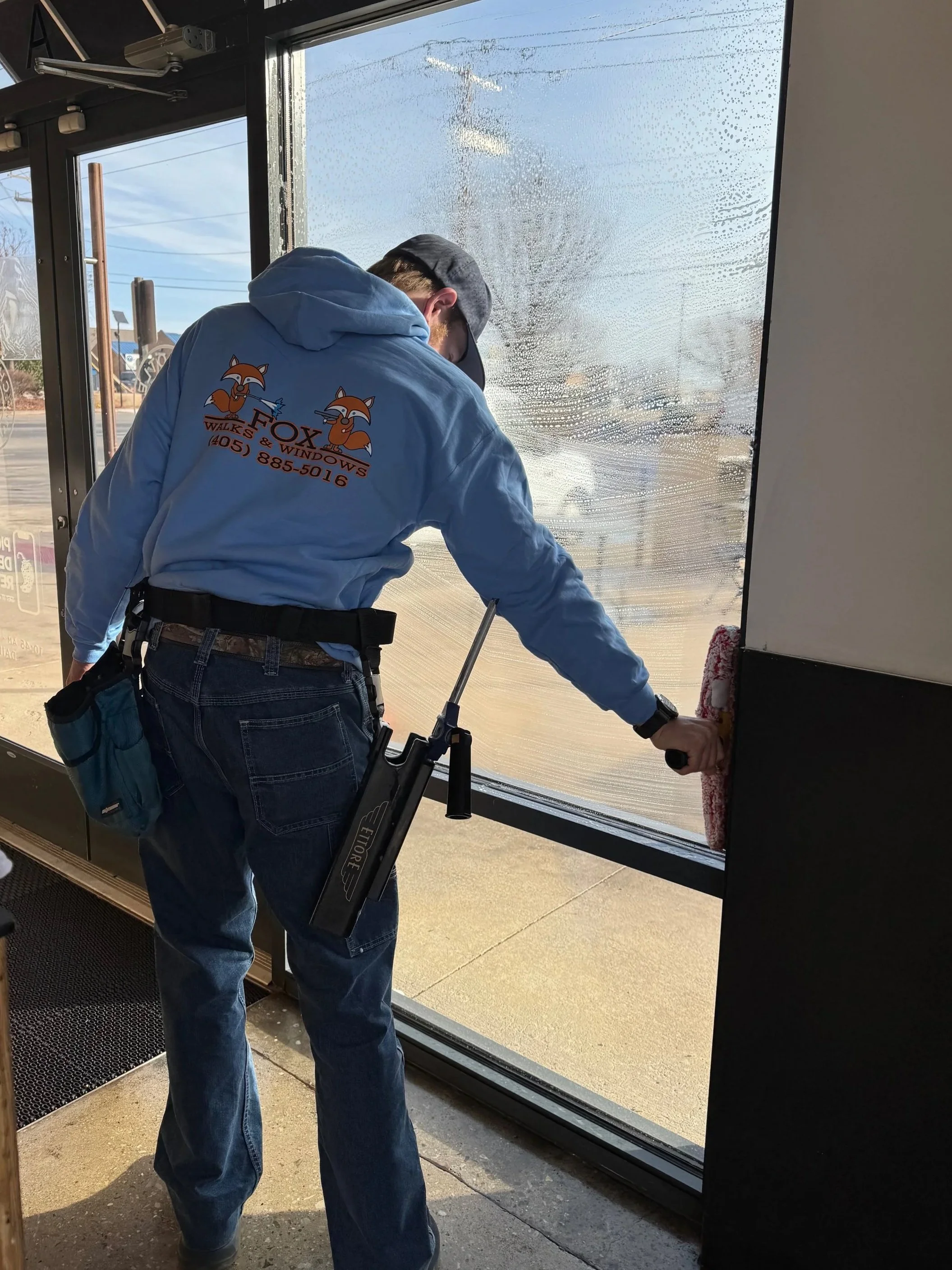 A worker cleaning the glass door with a red squeegee. The worker is wearing a blue hoodie with a fox logo and the text 'FOX WALKS & WINDOWS' on the back. Outside, the weather appears to be sunny and a parking lot is visible.