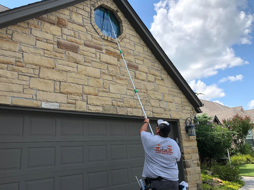 A man cleaning a small circular window on a house with a long-handled window cleaning tool.