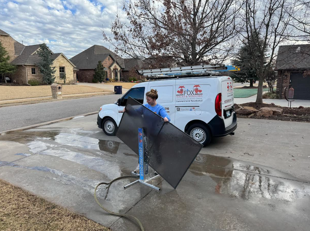 Woman cleaning a large window screen outdoors beside a white service van with ladders on top, parked on a residential driveway with houses and trees in the background.