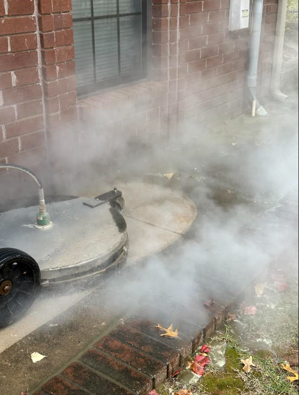 A sidewalk with steam rising from a ground cleanup machine next to a brick building at the corner of a sidewalk, with fallen autumn leaves scattered around.