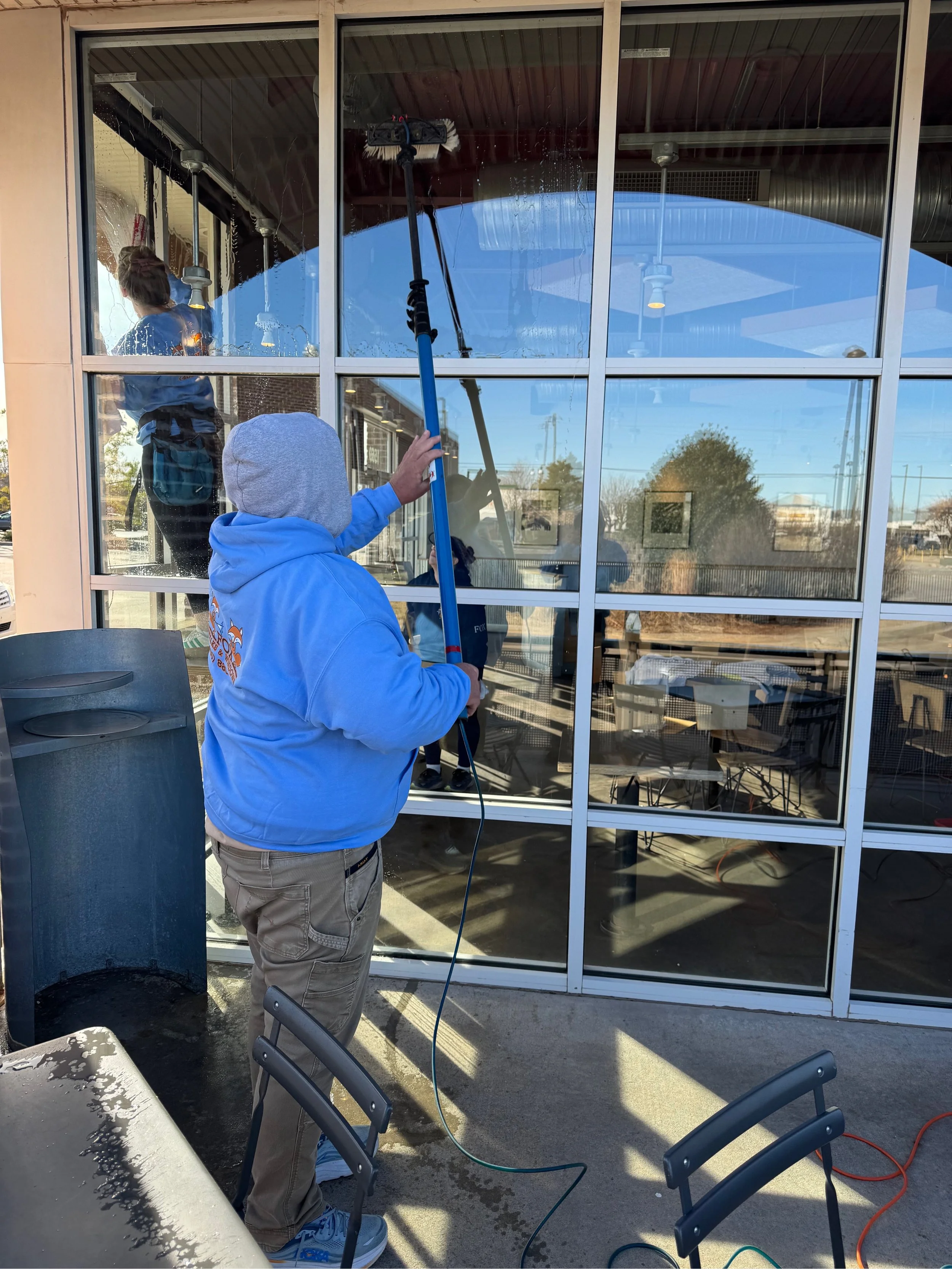 A person wearing a blue hoodie and gray hood is cleaning large glass windows with a long-handled squeegee outside a building.