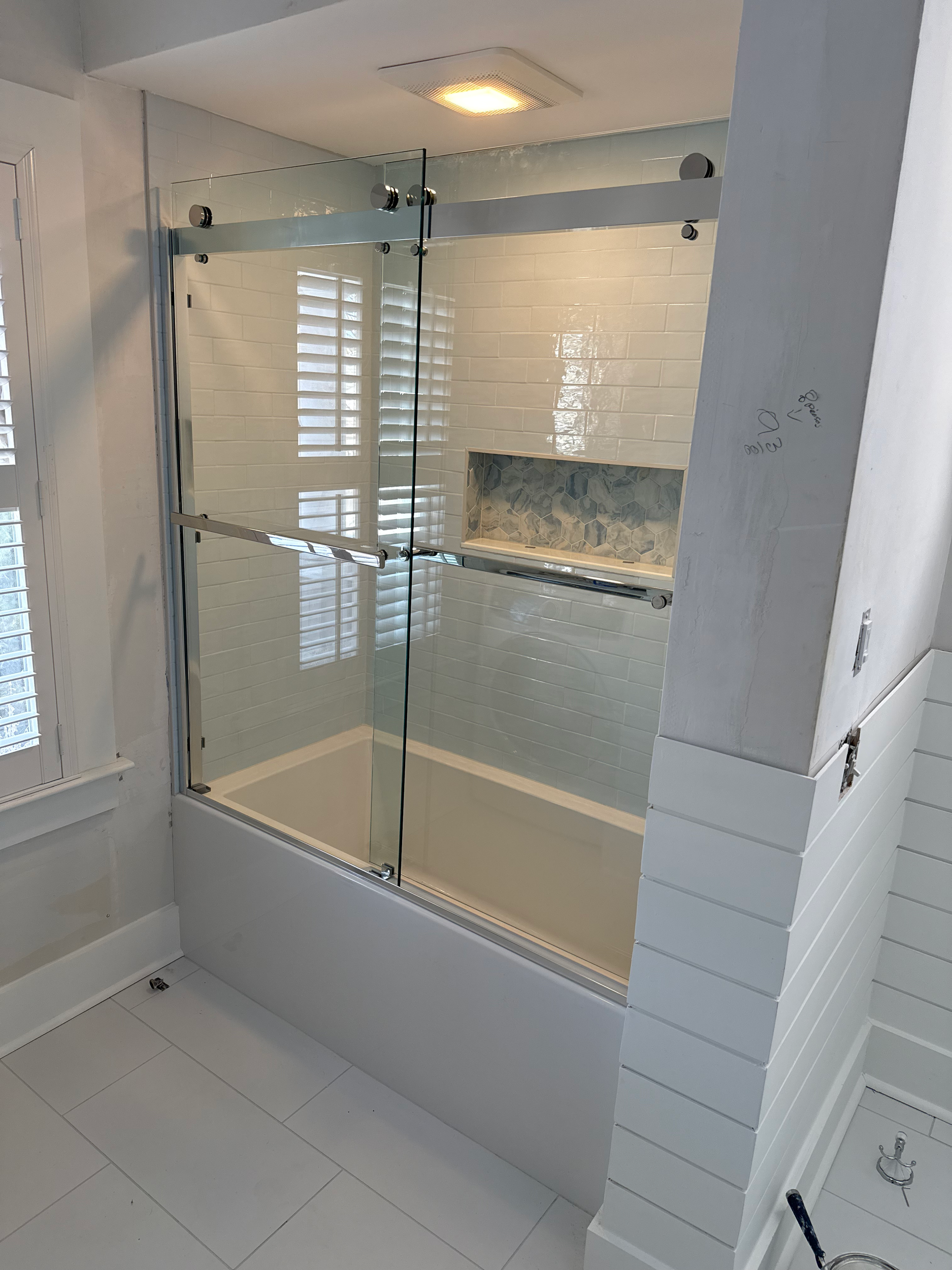 Modern bathroom with glass shower enclosure, white tiled walls, and an inset shower niche with hexagonal tiles. Light-colored floor tiles and a ceiling light are visible.