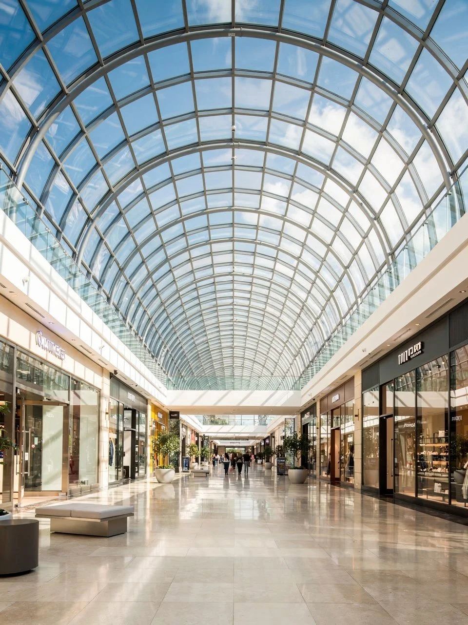 Interior of a spacious shopping mall with a glass arched ceiling, retail stores on both sides, and a few shoppers walking in the center.