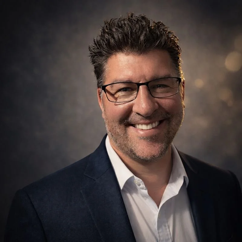A smiling man with glasses, dark hair, and a beard, wearing a dark suit jacket over a white shirt, with a blurred dark background.