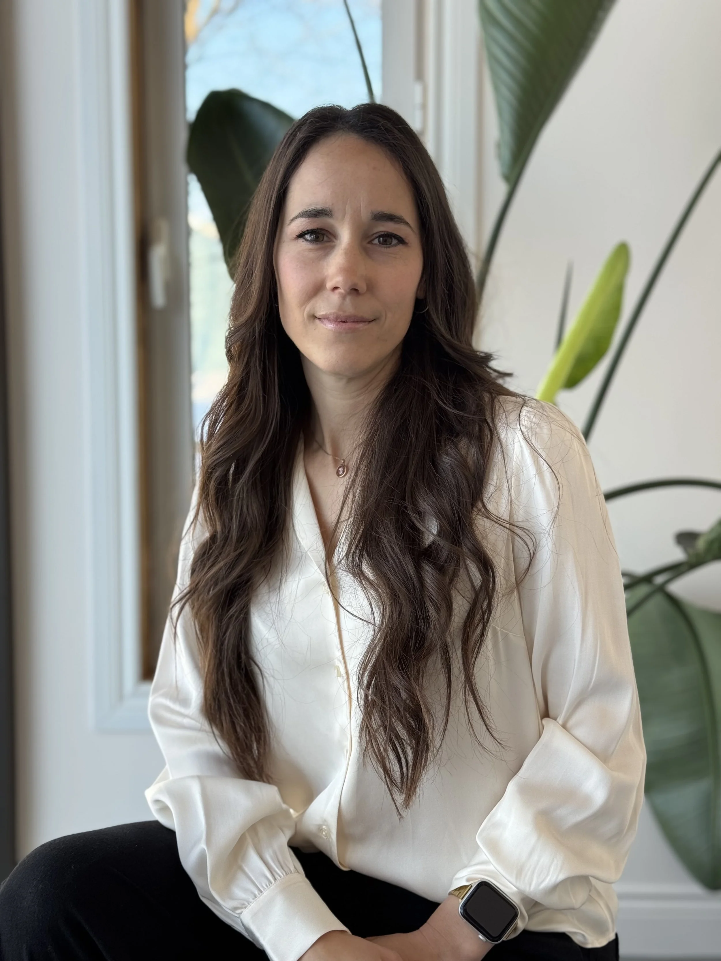 A woman with long wavy brown hair sitting indoors near large green plants, wearing a cream-colored silky blouse and a smartwatch on her left wrist.
