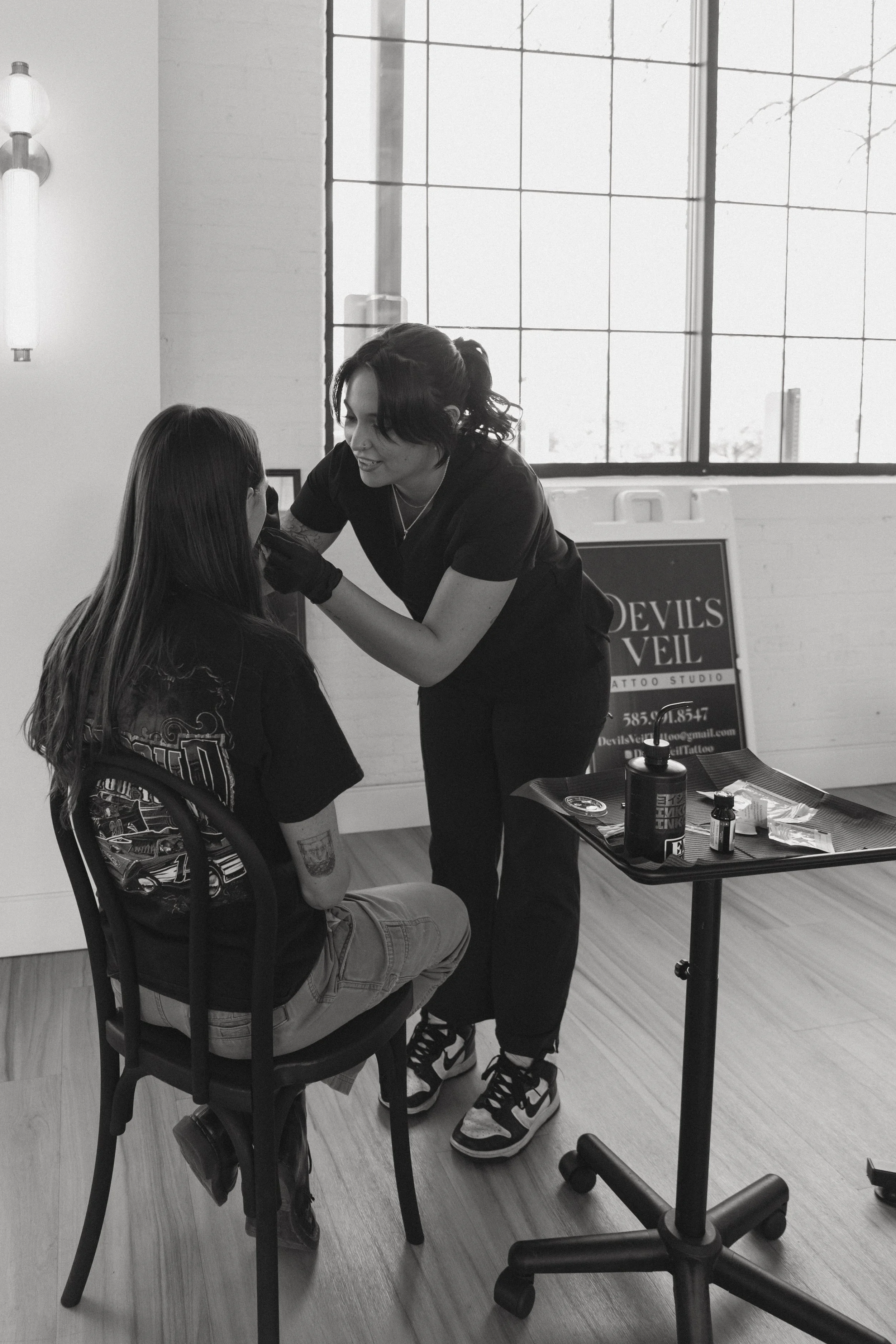 A tattoo artist is talking to a client seated in a chair, with tattoo equipment on a table nearby, inside a well-lit studio with large windows.