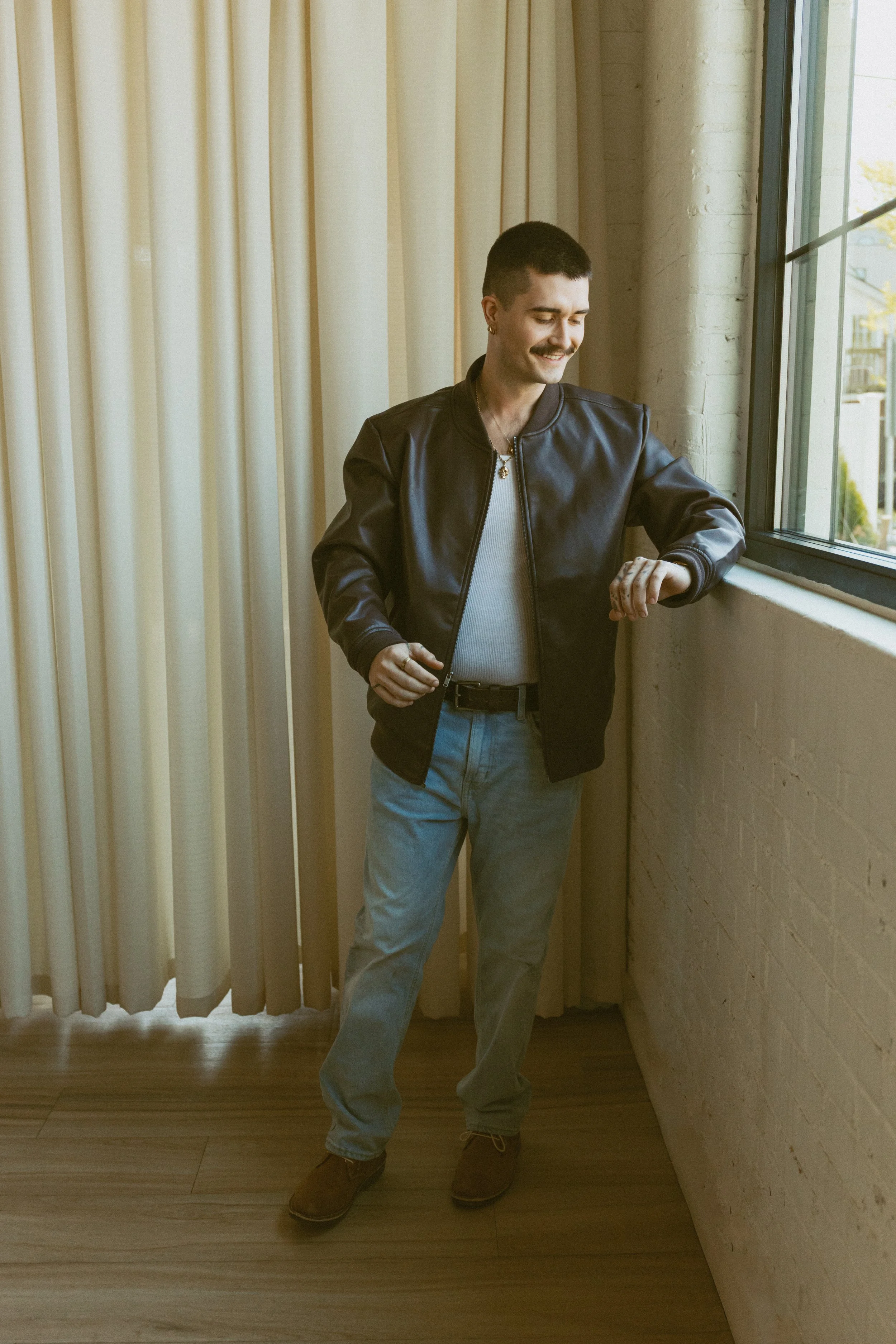 A young man with dark hair, a mustache, and earrings stands by a large window with beige curtains. He is wearing a black leather jacket, a light gray shirt, gray pants, and brown shoes. He is smiling and looking at his watch, with one hand slightly r