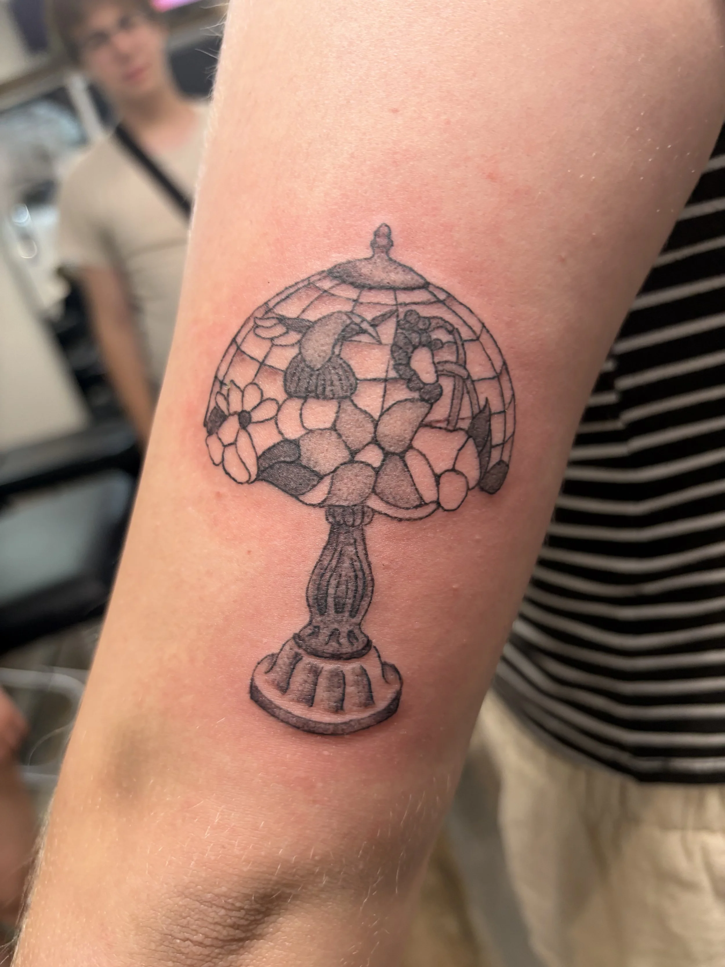 A tattoo of a vintage table fan on a person's upper arm. The tattoo is outlined in black ink and depicts the fan's blades, protective cage, base, and handle. A person with curly hair and glasses can be seen blurred in the background.