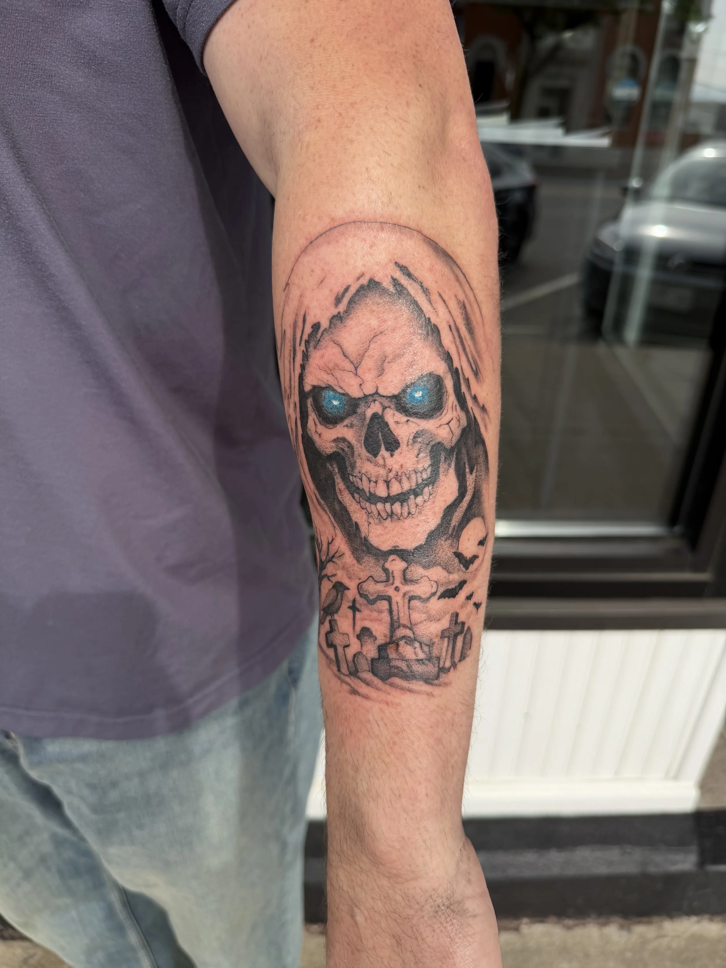 Tattoo of a skull with glowing blue eyes on the person's forearm, with crosses and gravestones below it.