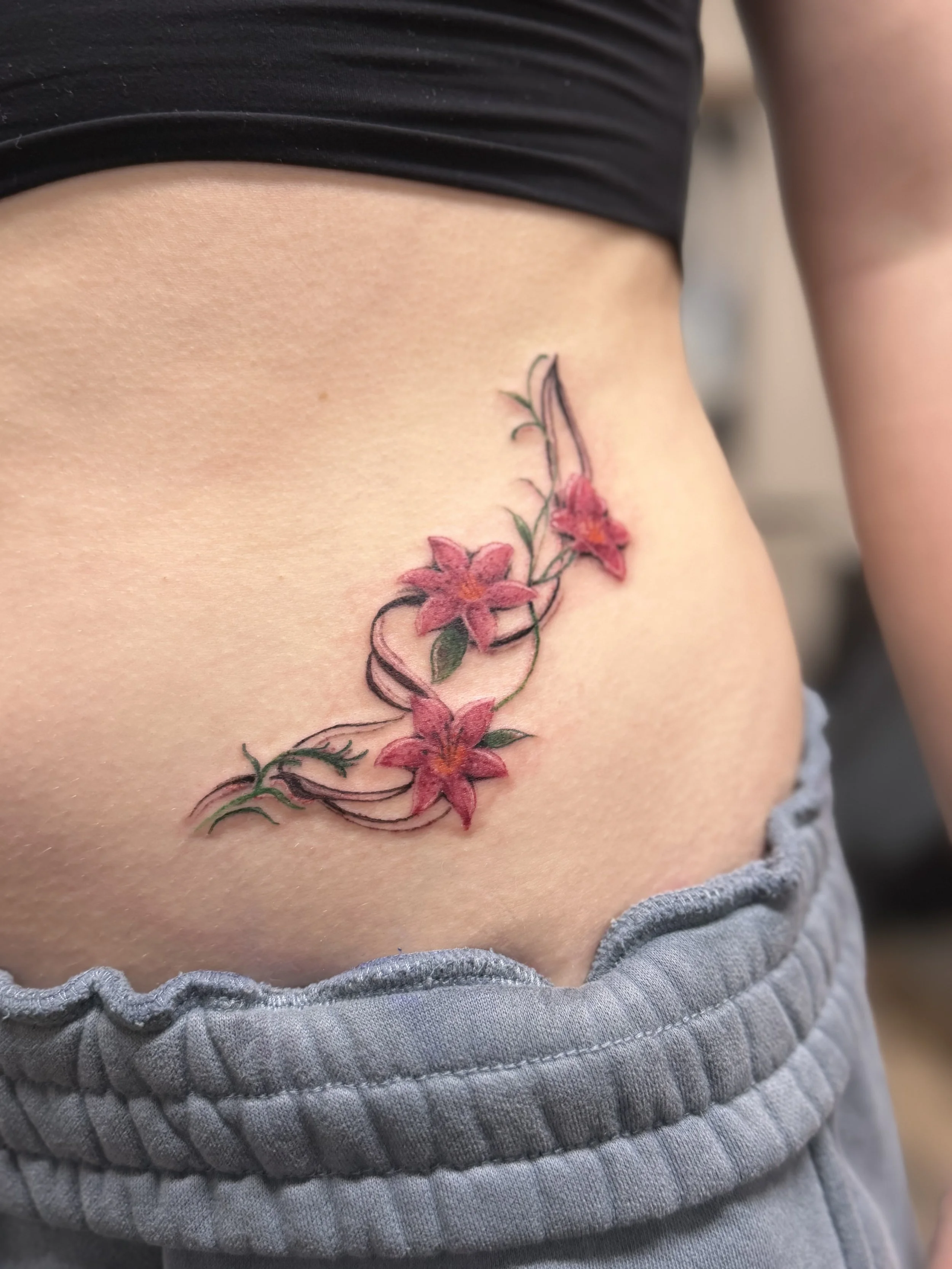 Close-up of a person's side torso featuring a colorful floral tattoo with pink flowers and green leaves on the ribcage, with the person wearing a black crop top and gray sweatpants.
