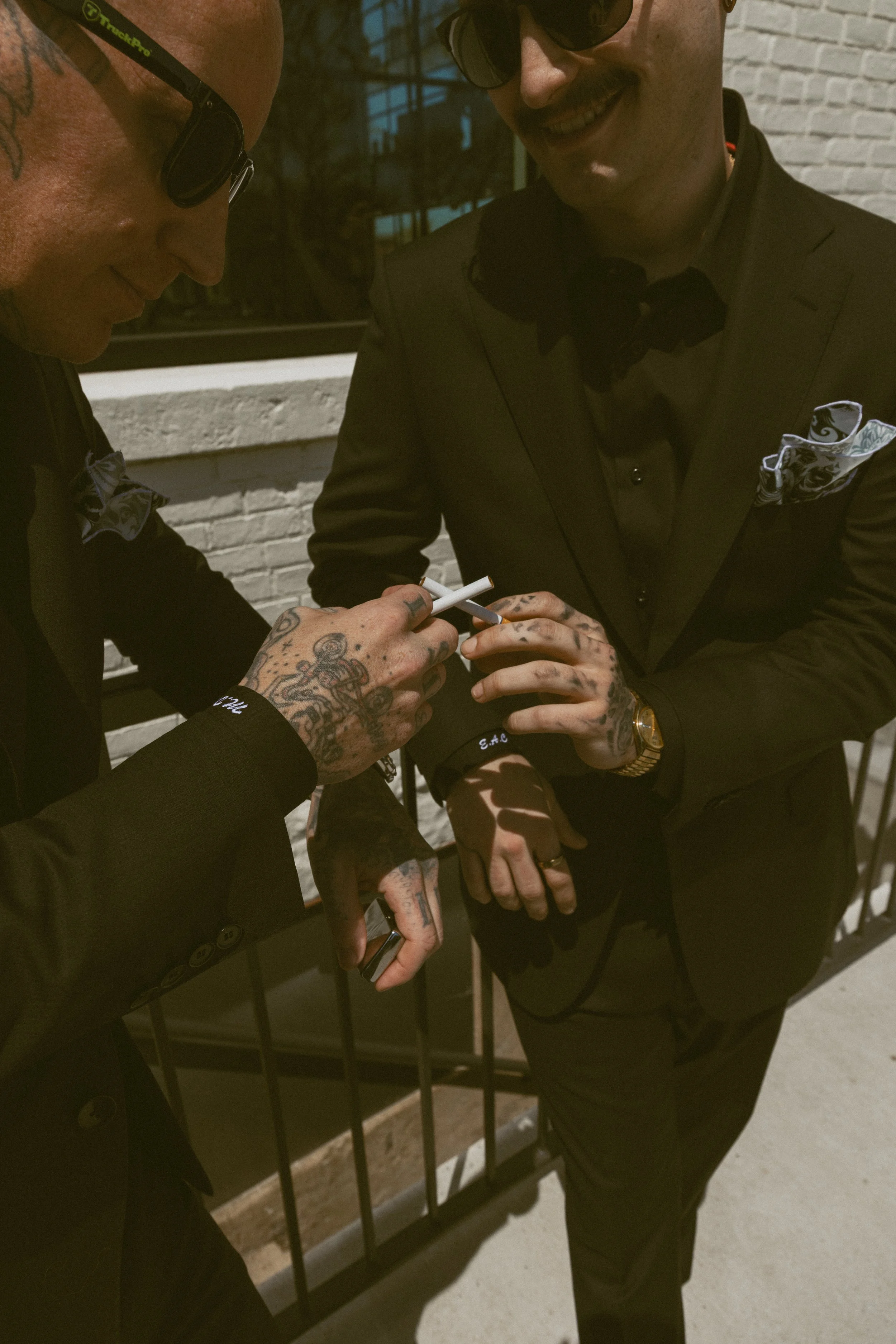 Two men in suits with bow ties and pocket squares are smiling while signing an autograph. One has tattoos on his hands and is wearing glasses, the other has a mustache and is holding his hands together. They are standing in front of a brick wall and 