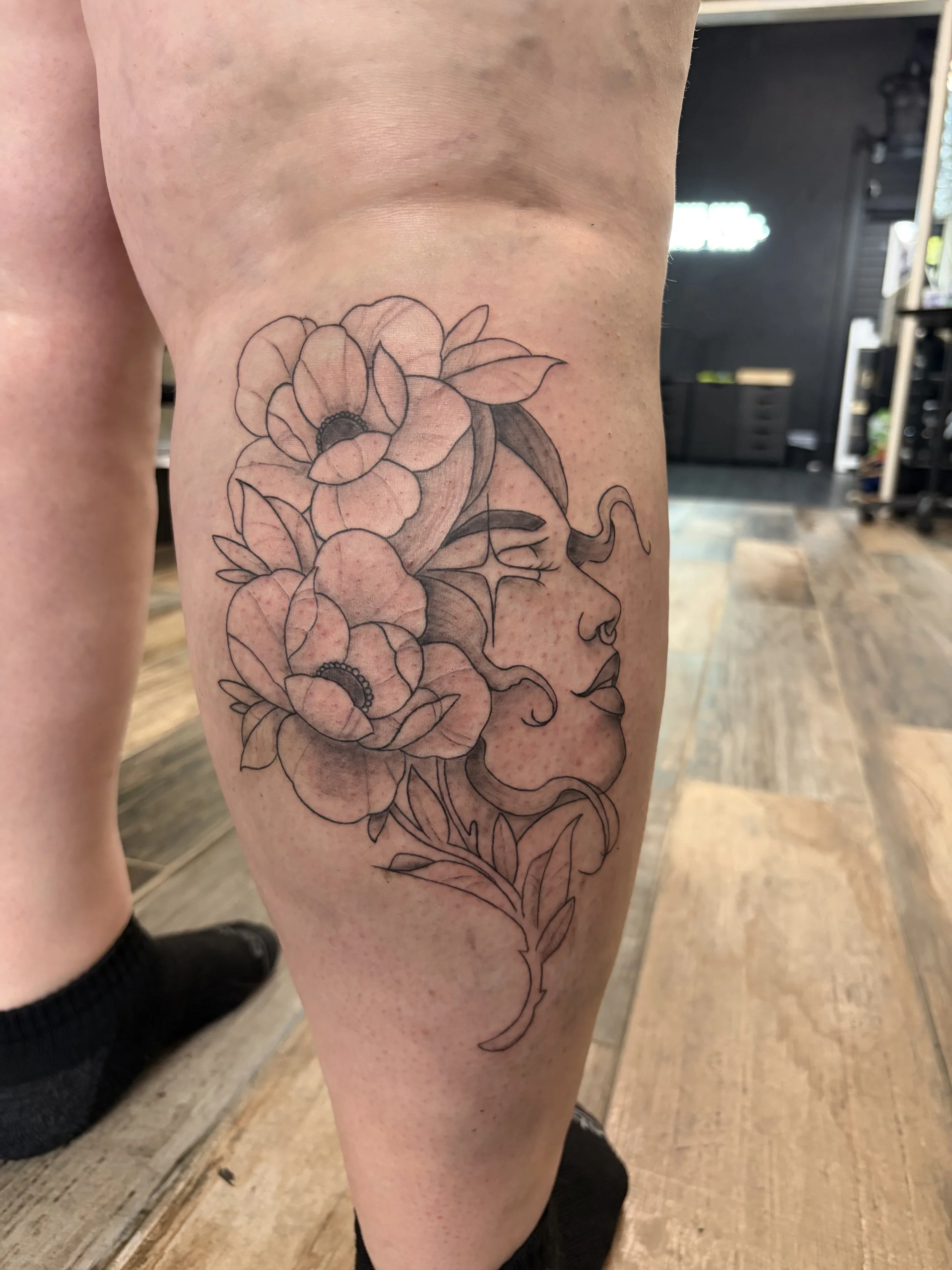 A black and white line art tattoo of flowers and a woman's profile on someone's thigh.