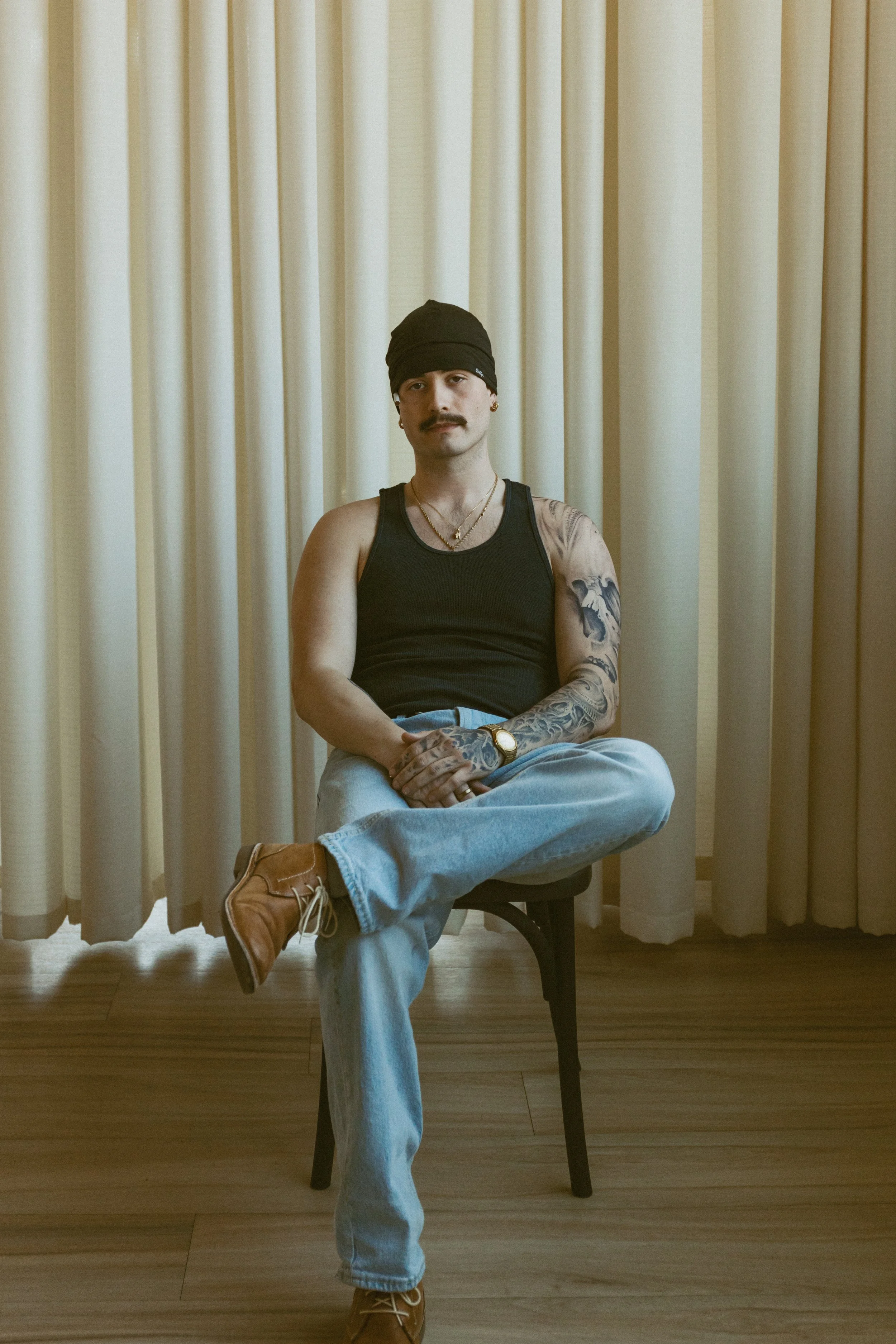 A man with a mustache and tattoos on his arms, wearing a black bandana, black tank top, light blue jeans, tan boots, and gold jewelry, sitting cross-legged on a chair in front of beige curtains.