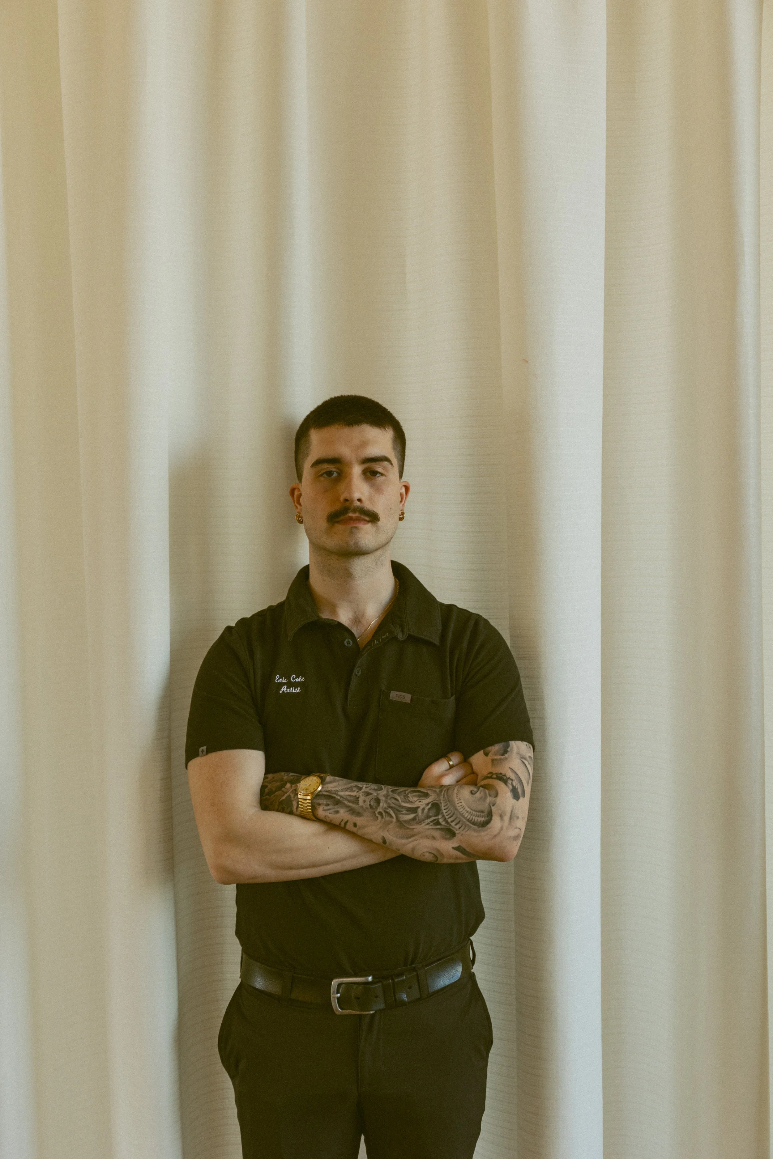 A man with short dark hair, a mustache, and earrings stands with his arms crossed in front of a beige curtain. He is wearing a black polo shirt with a name tag that reads "Eric Cale Artist" and has tattoos on his forearms.
