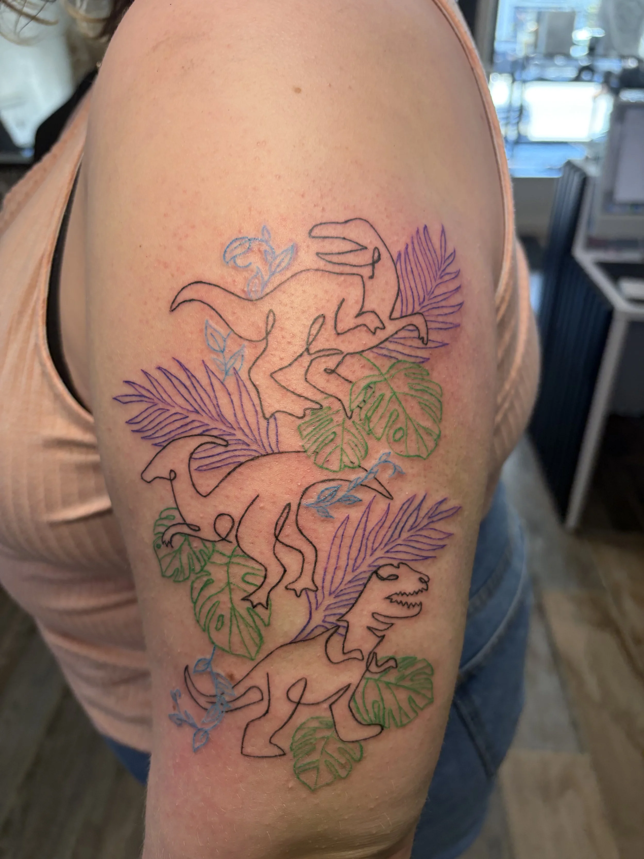 A tattoo of a dinosaur surrounded by tropical leaves on a person's upper arm.