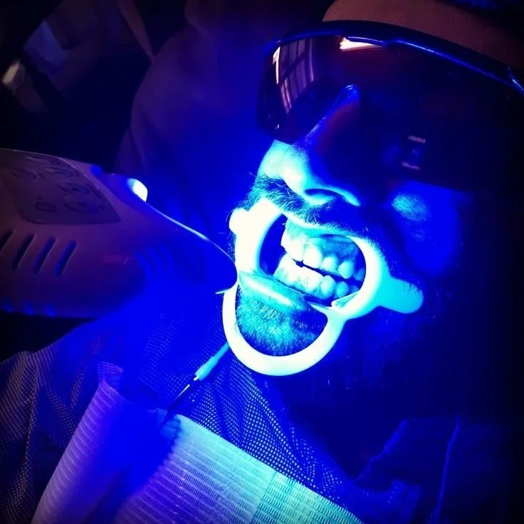 A person wearing protective glasses and a face shield is smiling with their mouth open, showing their teeth, under a blue light.