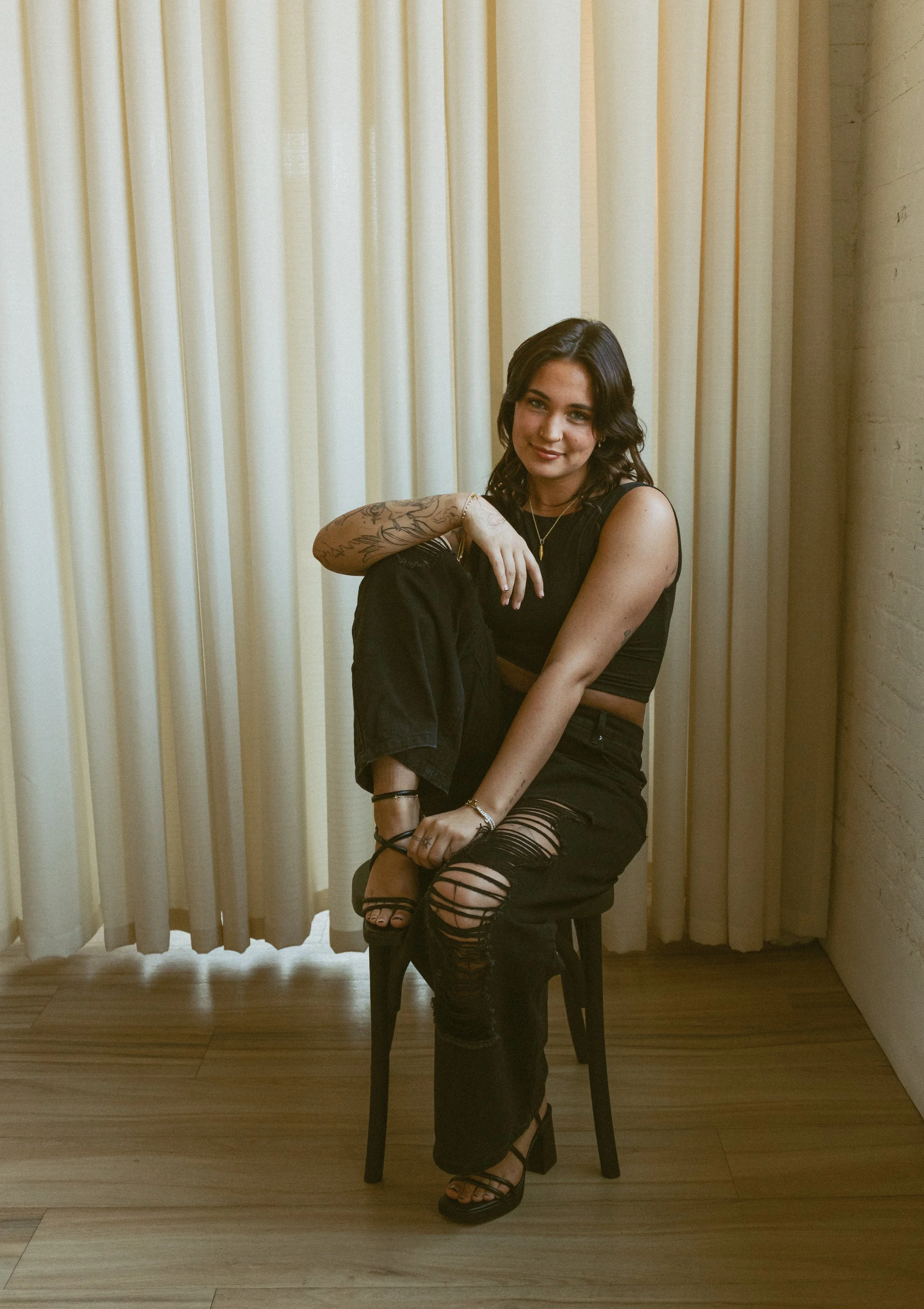 A woman with dark wavy hair sitting on a chair in front of cream-colored curtains. She is wearing a sleeveless black top, ripped black pants, and black high-heeled sandals. She is smiling and has tattoos on her left arm.