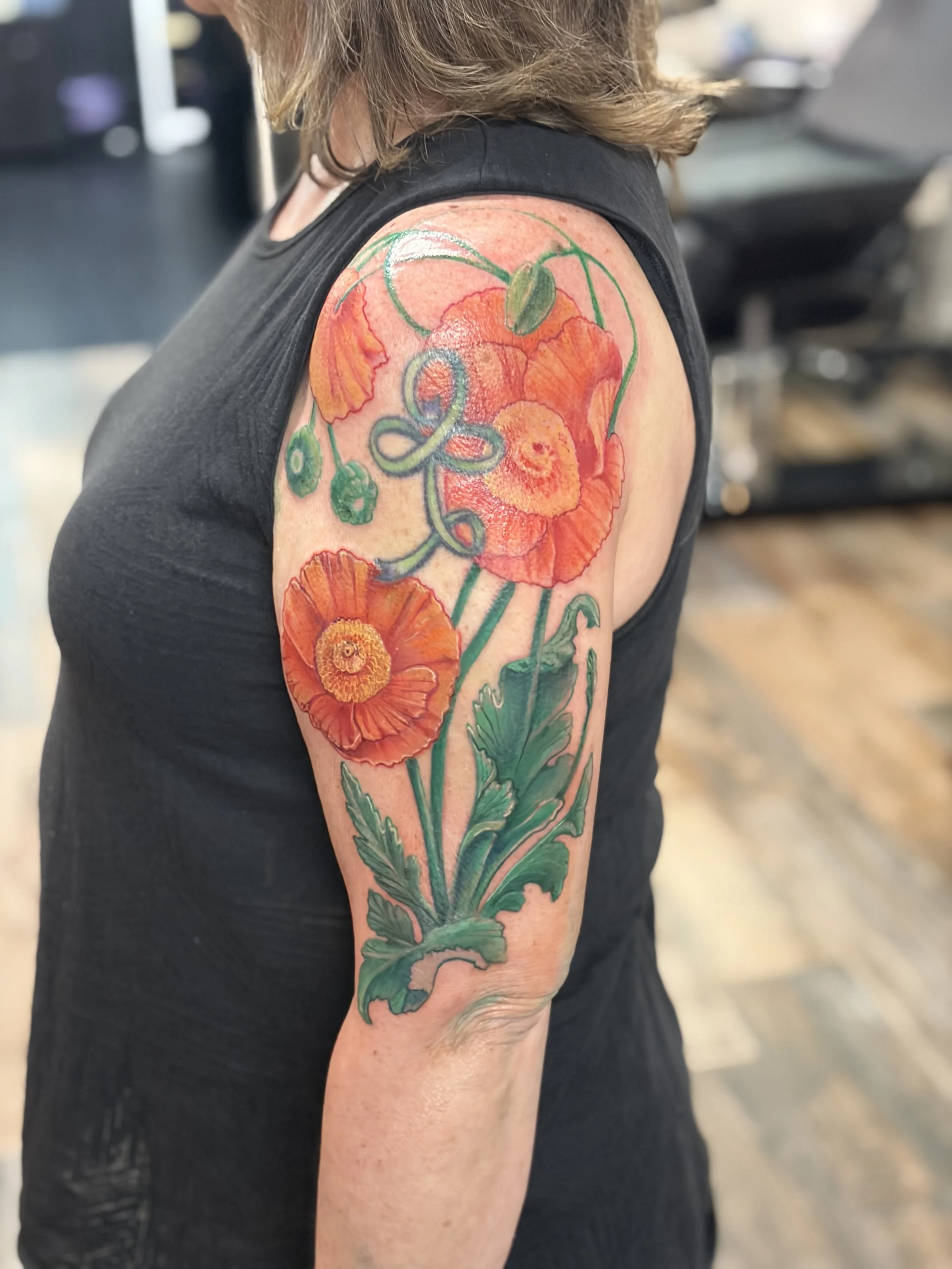 Colorful floral tattoo on person's upper arm and shoulder featuring orange poppies and green leaves.