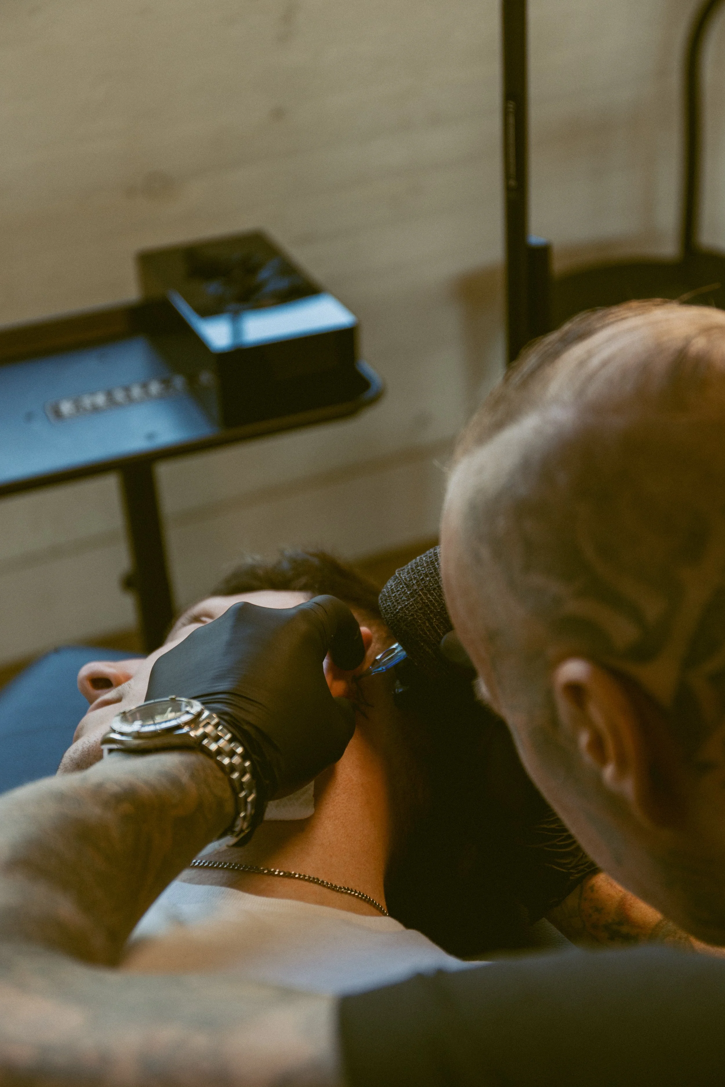 A person with a bald head and tattoos receives a tattoo on their back from an artist wearing black gloves. The scene takes place indoors, with a beige wall and furniture visible in the background.
