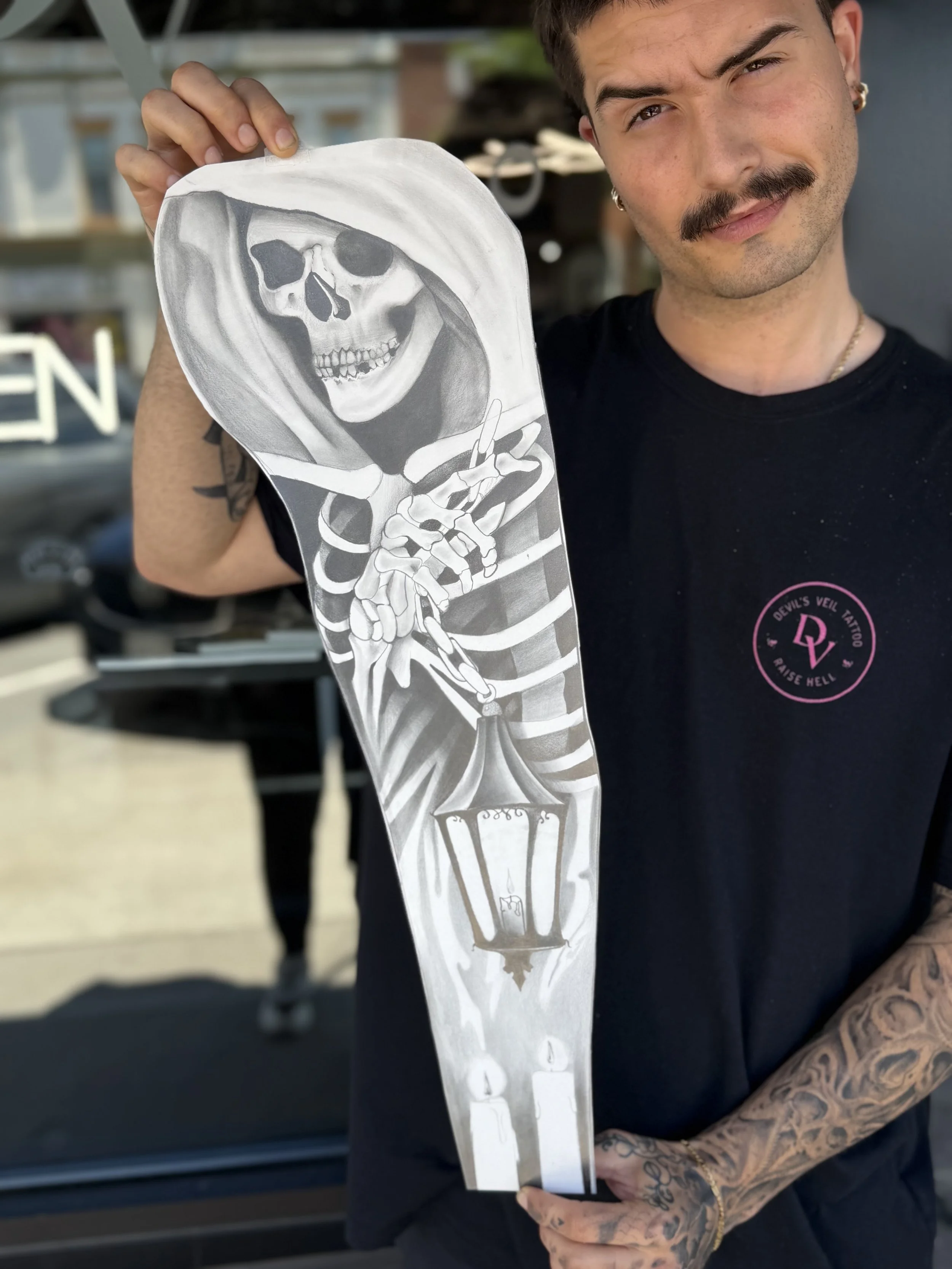 Man with tattoos holding a paper cutout of a skeleton in a hooded cloak with a broad smile and selective shading.