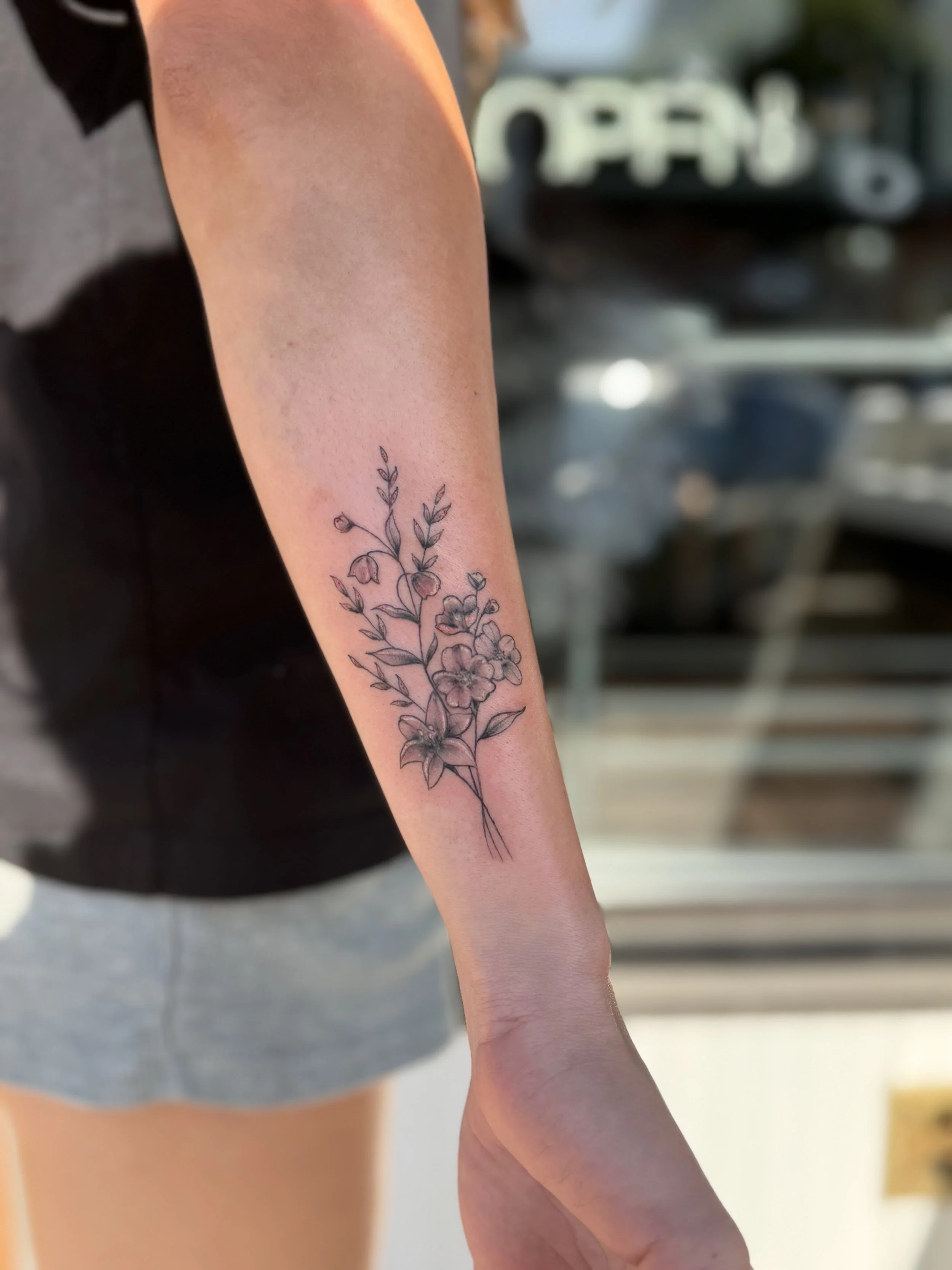 A close-up photo of a person's forearm with a black outline tattoo of a flowering plant. The person is standing outdoors near a coffee shop or similar setting, with a sign that says 'OPEN' in the background.