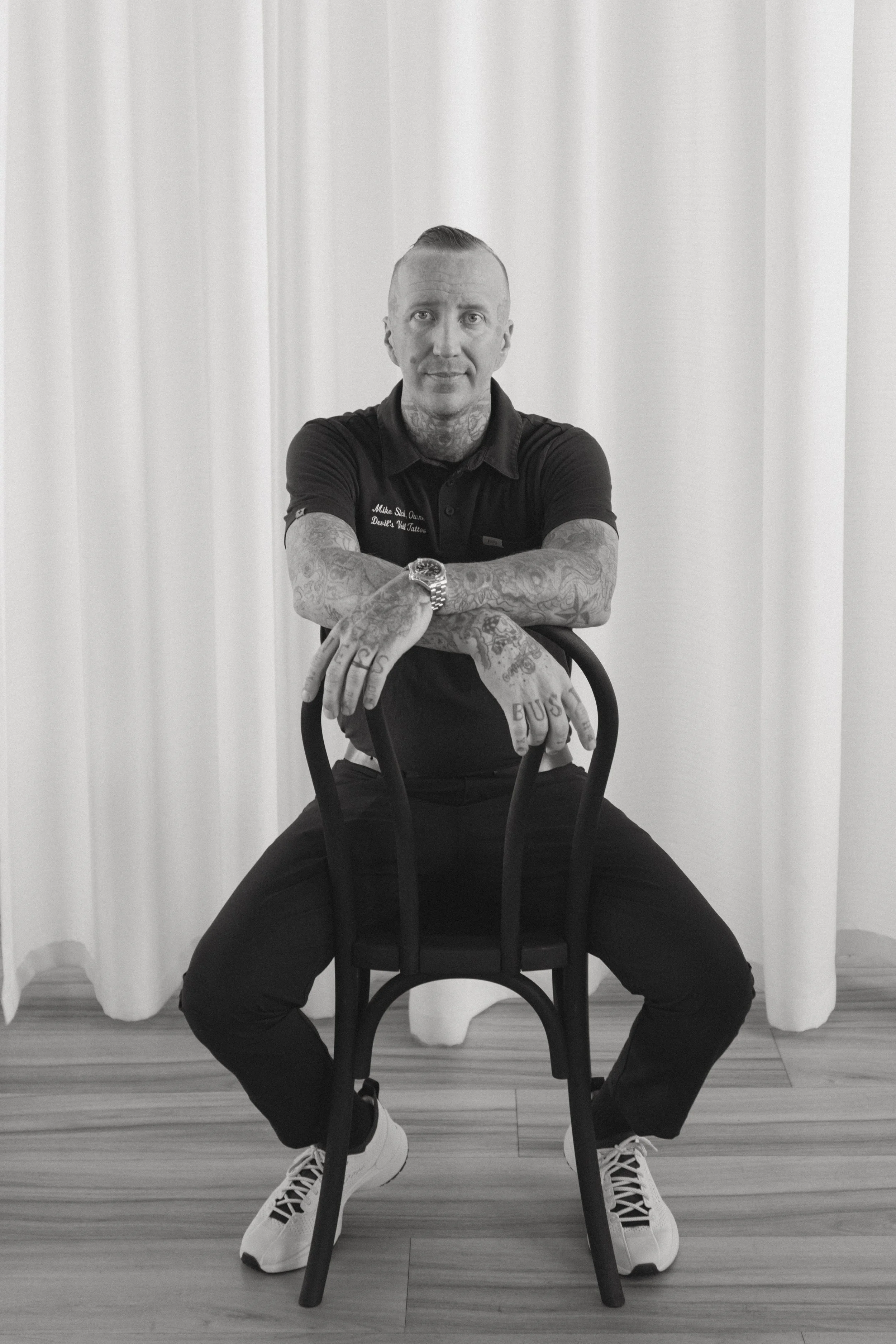 A man with tattoos and short hair sitting on a chair with his arms crossed, wearing black pants, white sneakers, and a dark polo shirt, in front of plain white curtains.