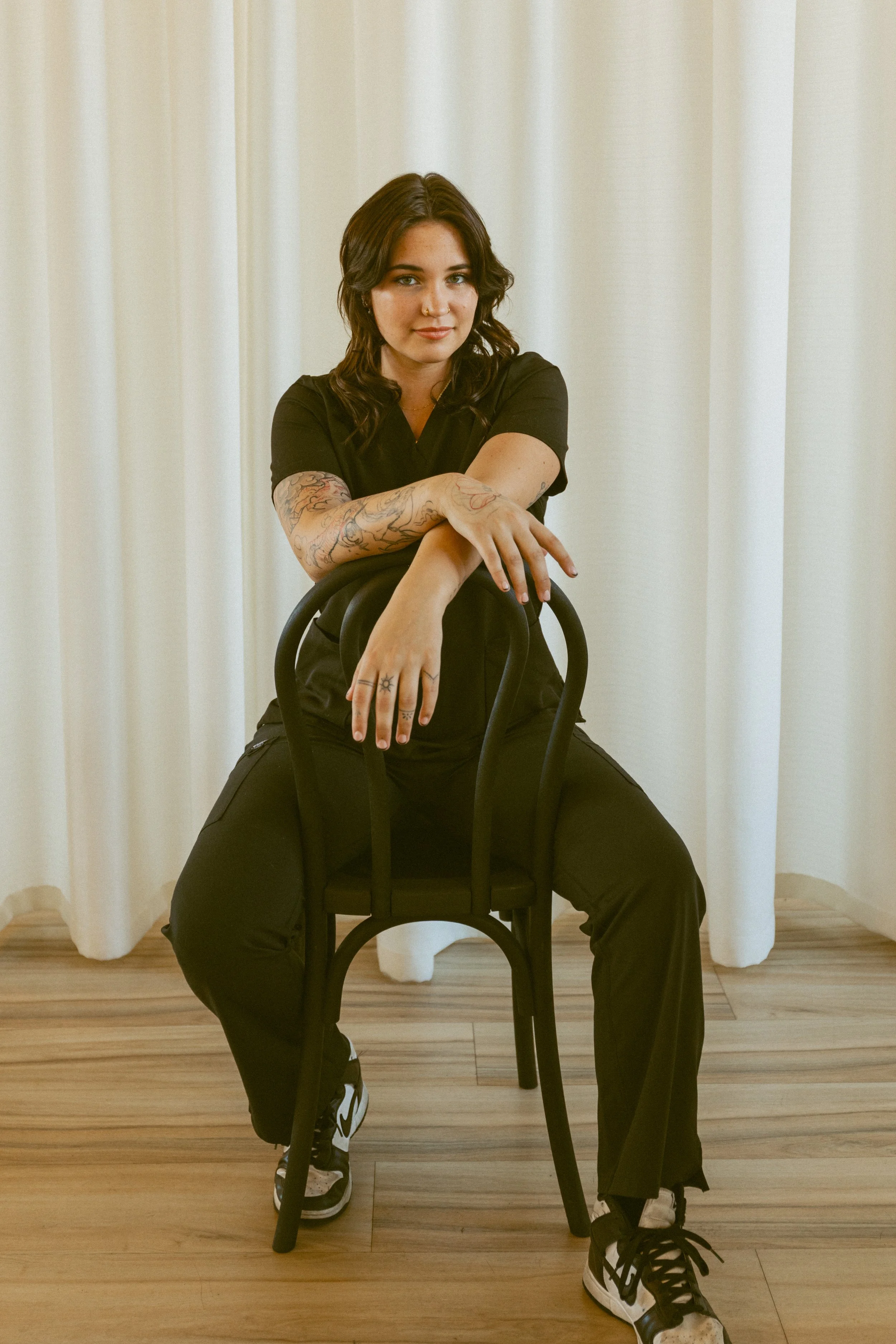 A young woman with dark brown wavy hair, wearing a black shirt and black pants, sitting on a black chair with her arms crossed and resting on the back of a second black chair, in front of cream-colored curtains and a wooden floor.