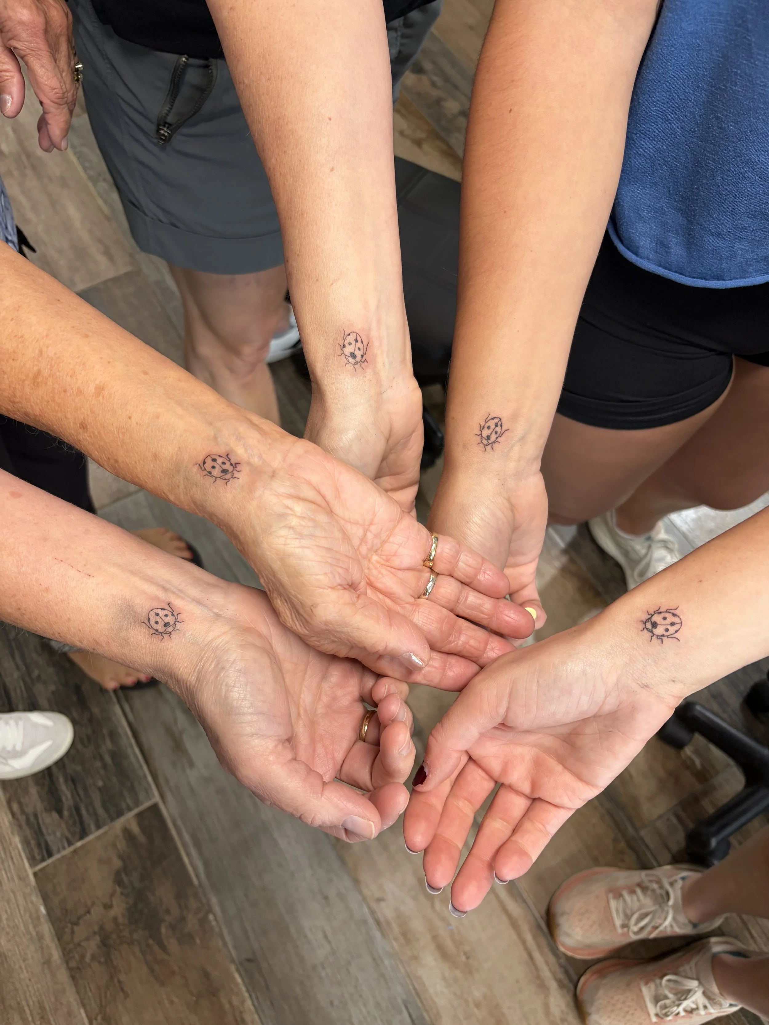 Multiple hands joined together in a circle, each with a small ladybug tattoo on the inner wrist or forearm.