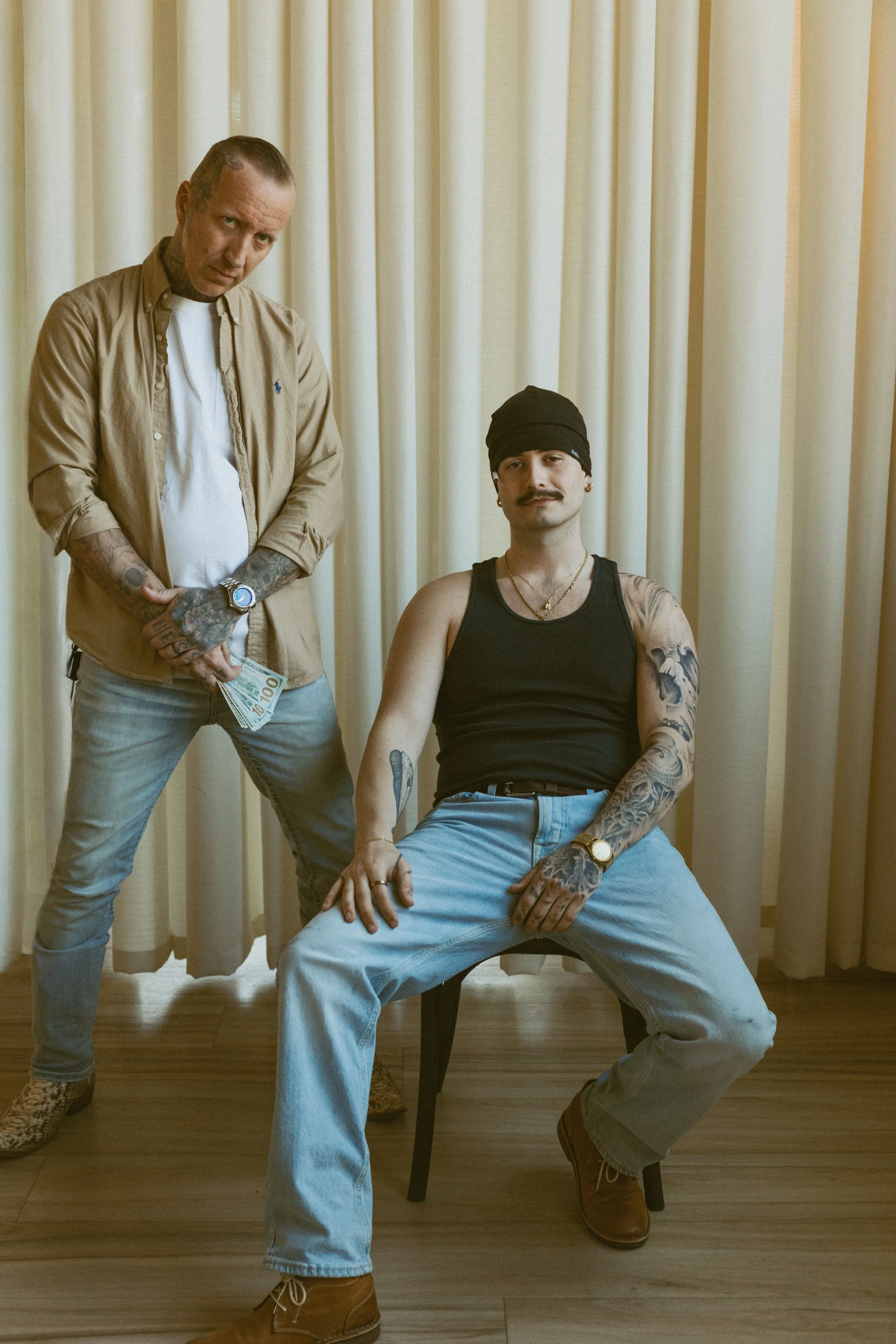 Two men with tattoos, one standing holding money and one sitting with a hand on his thigh, in a room with yellow curtains.