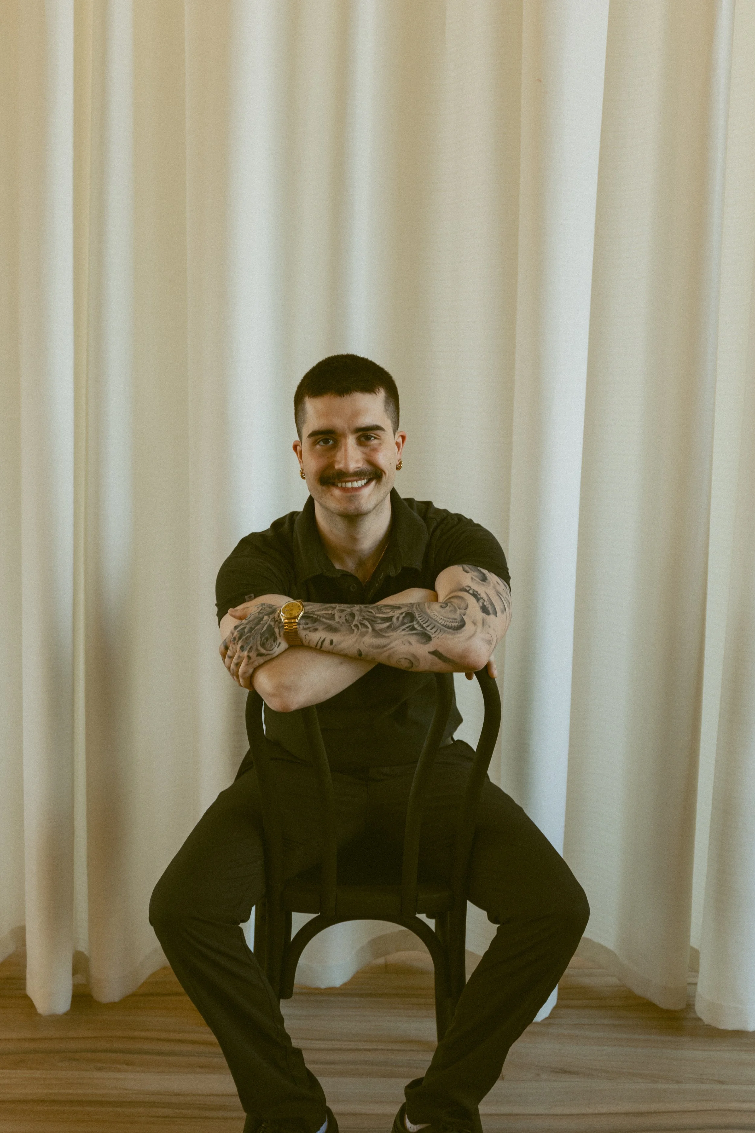 A man sits on a black chair in front of a beige curtain. He has short dark hair, tattoos on his arms, and is smiling with his arms crossed on his lap. He's wearing a black shirt, black pants, and glasses.