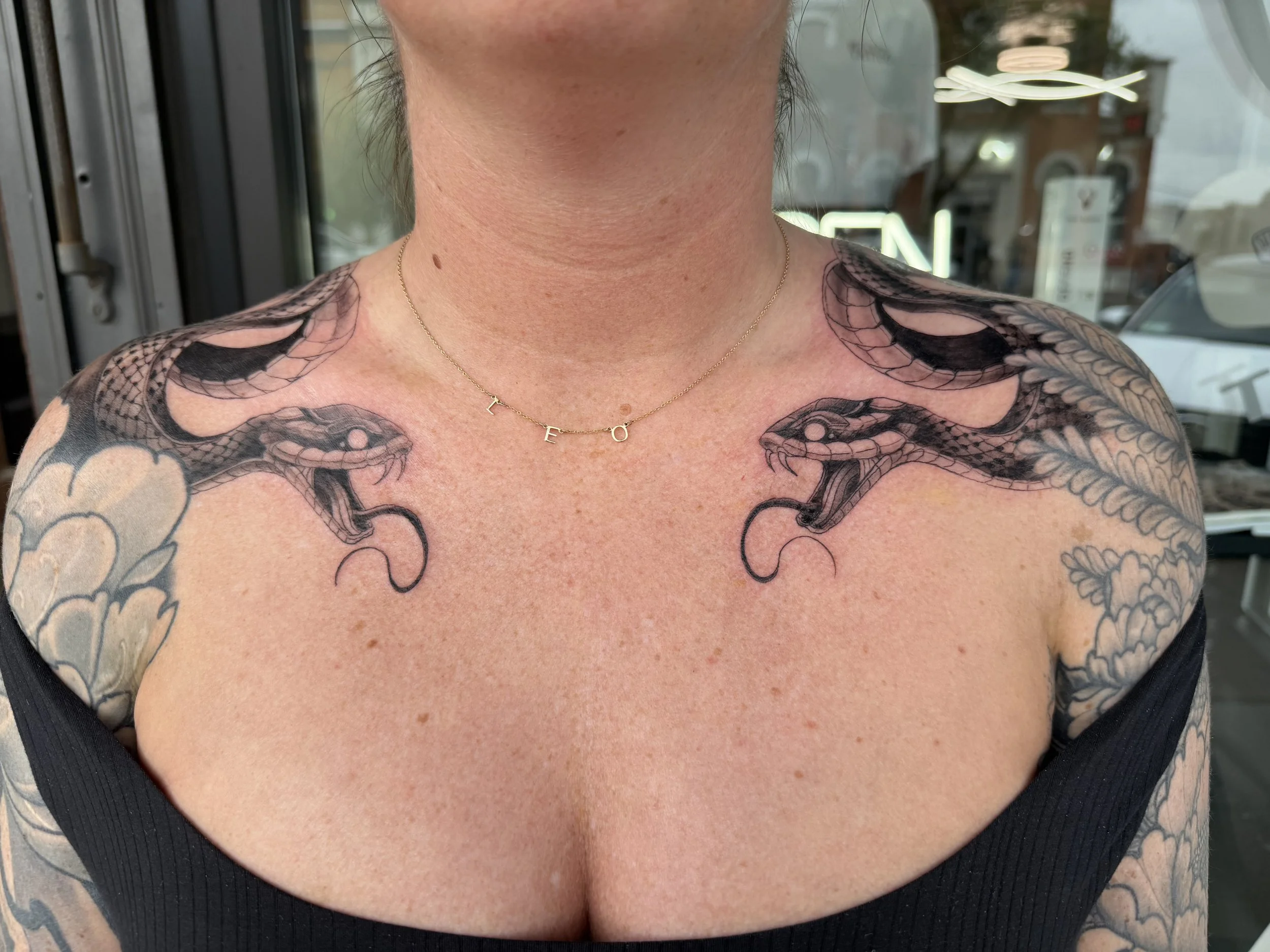 Close-up of a person's neck and upper chest with tattoos and a gold necklace with the letters 'L', 'E', and 'O' on it.