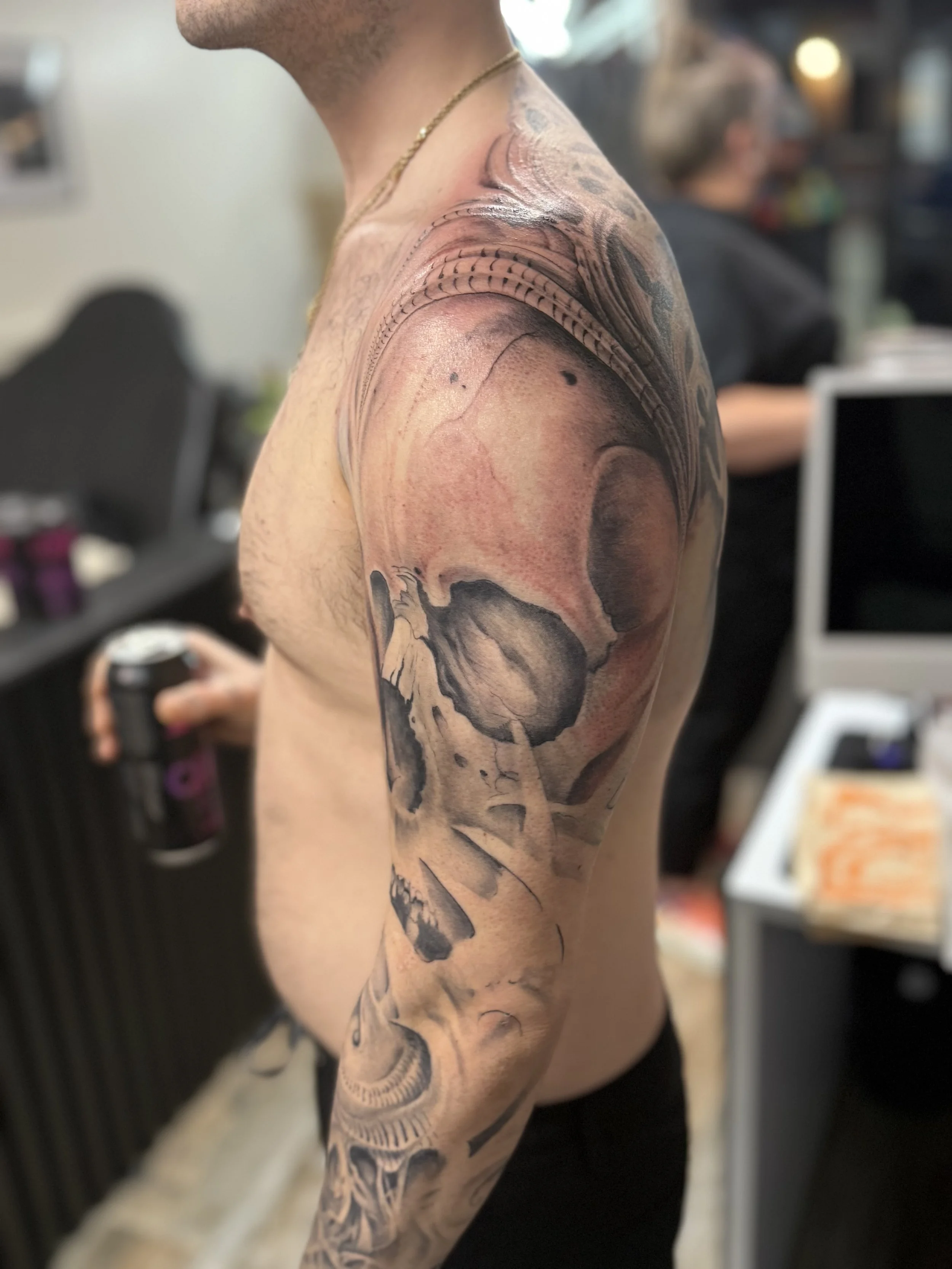 A person with a large black and gray tattoo sleeve on their right arm, standing indoors in a casual setting.
