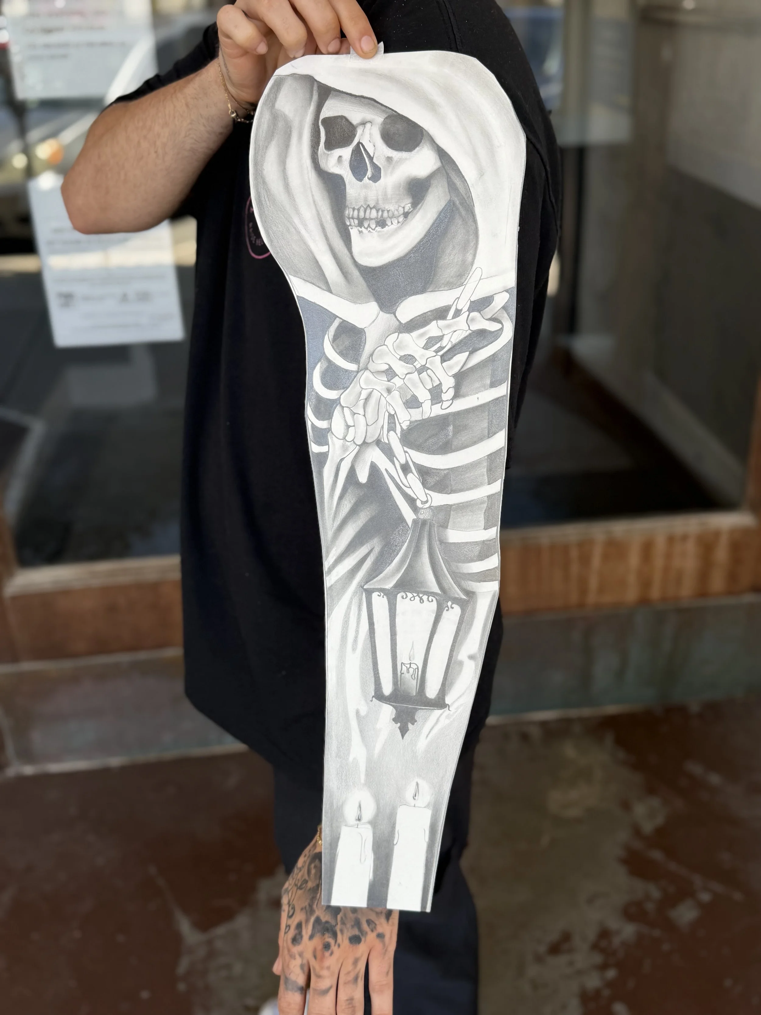 A person holding a paper drawing of a skeleton with a hood and smoking a cigarette, with candles at the bottom.