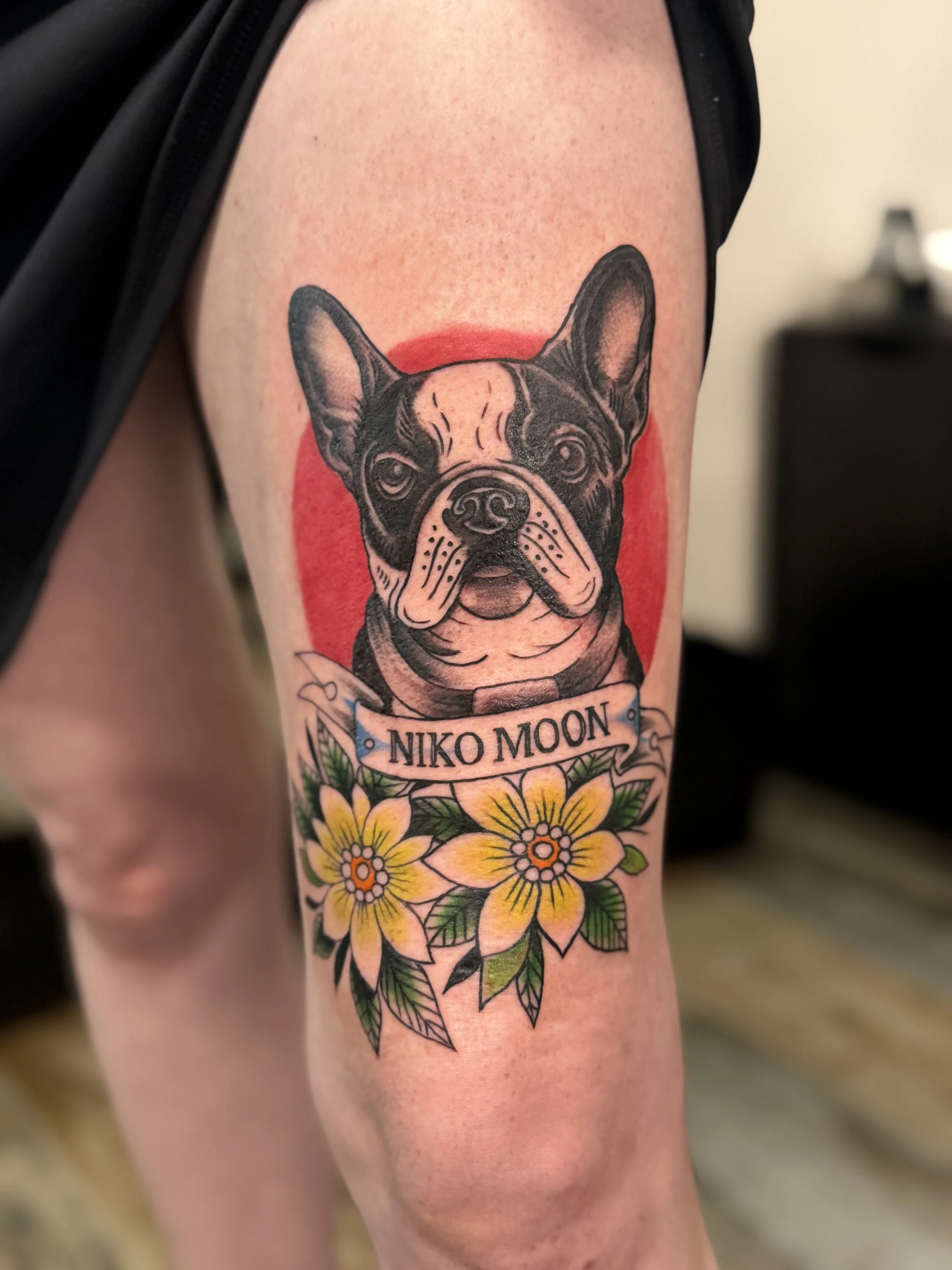 Tattoo of a French Bulldog with the name Niko Moon on the arm, surrounded by colorful flowers.