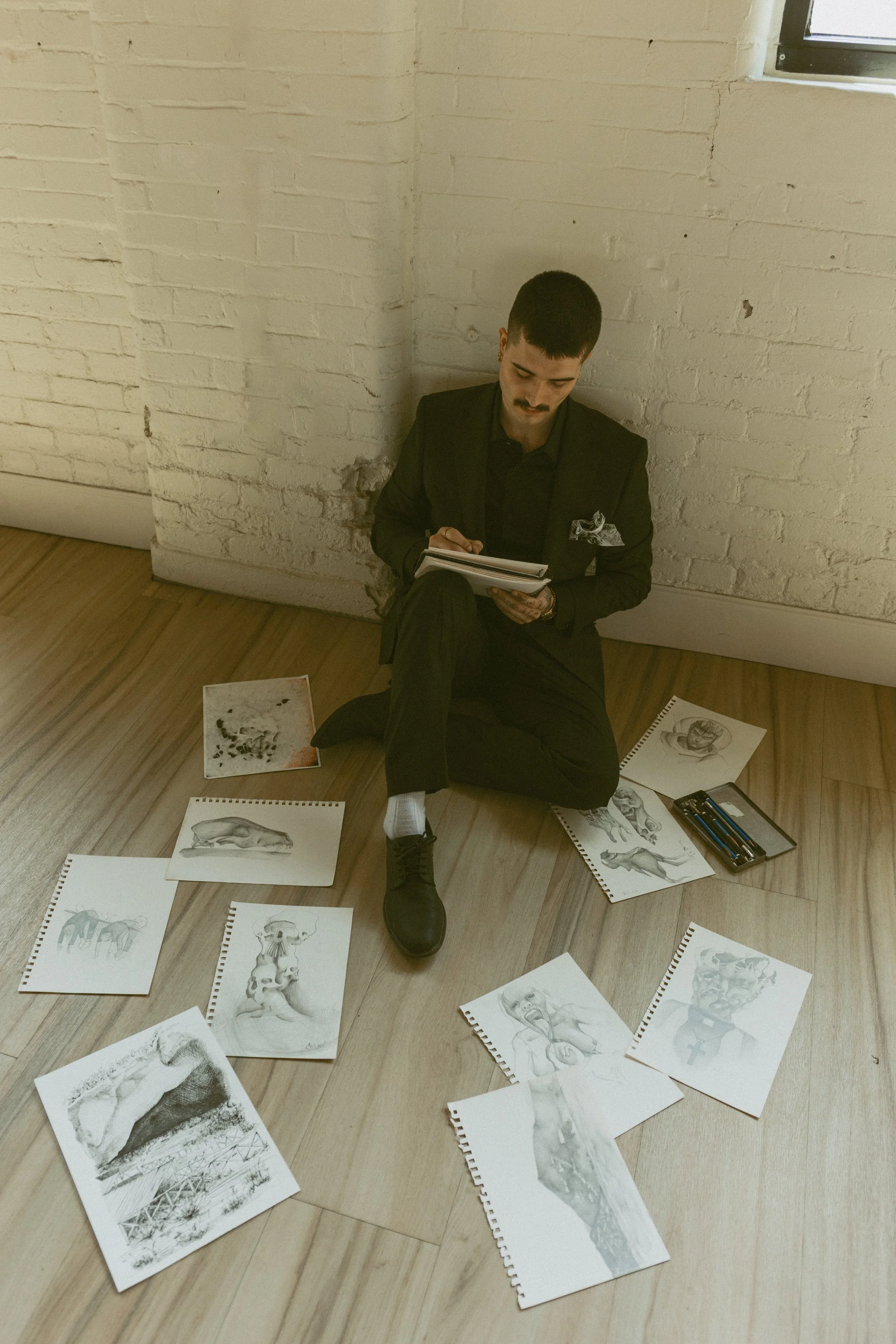 A man dressed in black sits on the floor against a white brick wall, surrounded by detailed black-and-white sketches of human and animal anatomy. He is looking at a clipboard, with various drawings scattered around him on the hardwood floor.