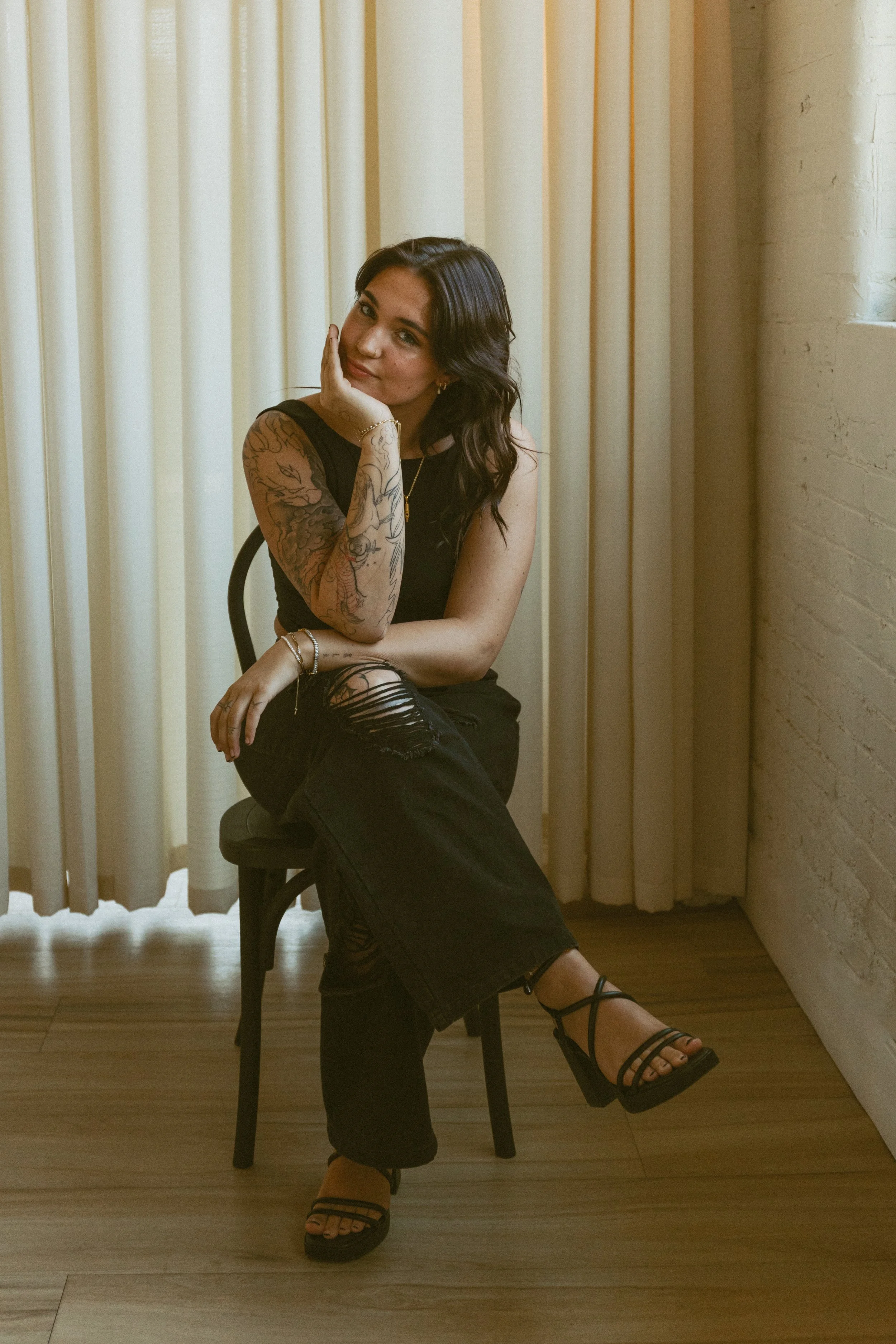 A woman with dark wavy hair sitting on a chair with one leg crossed over the other, resting her chin on her hand, wearing a sleeveless black top, ripped black jeans, and black high-heeled sandals, in front of cream-colored curtains and a white brick 
