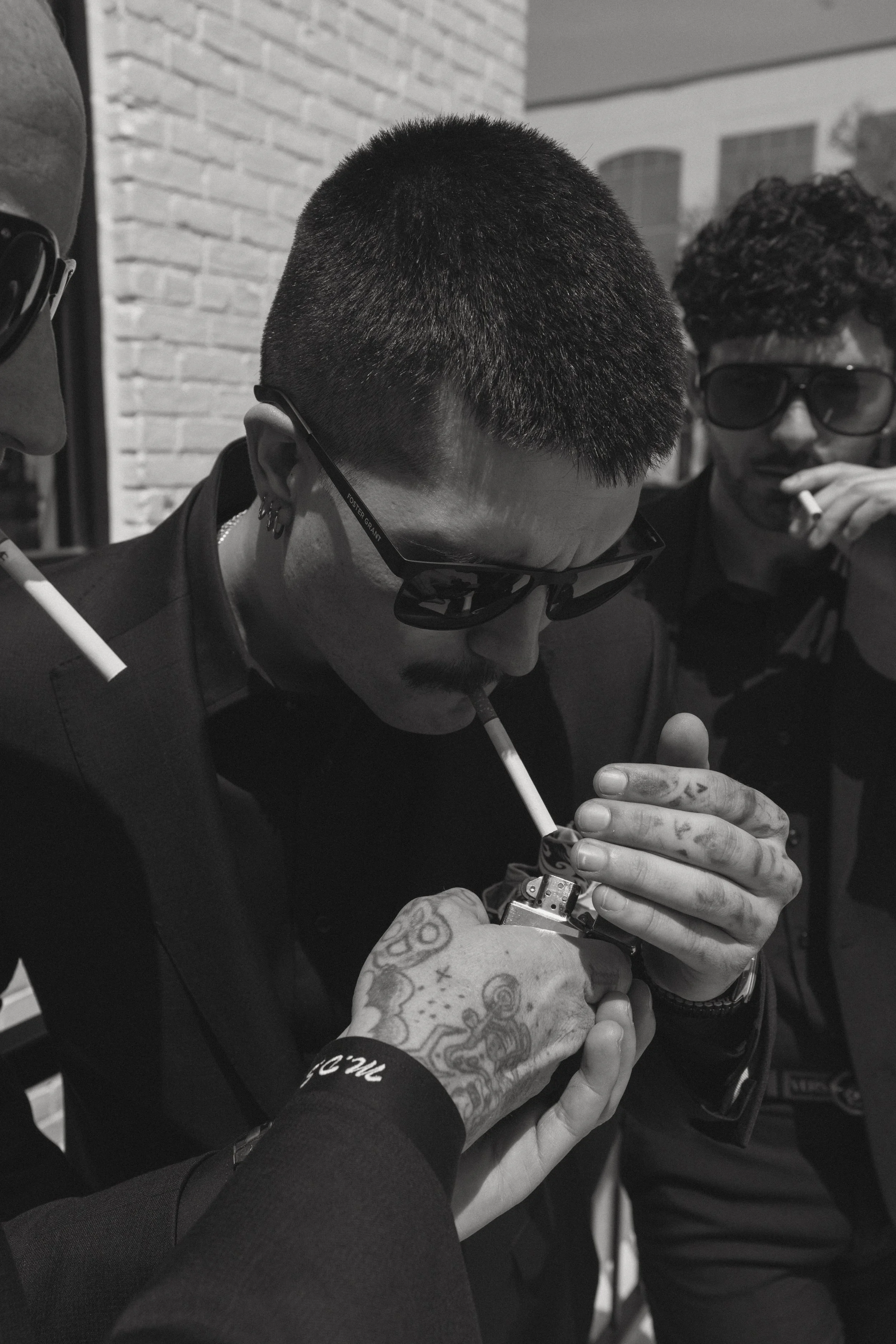 Two men with tattoos and sunglasses are focused on a tattoo machine, working together on a tattoo.