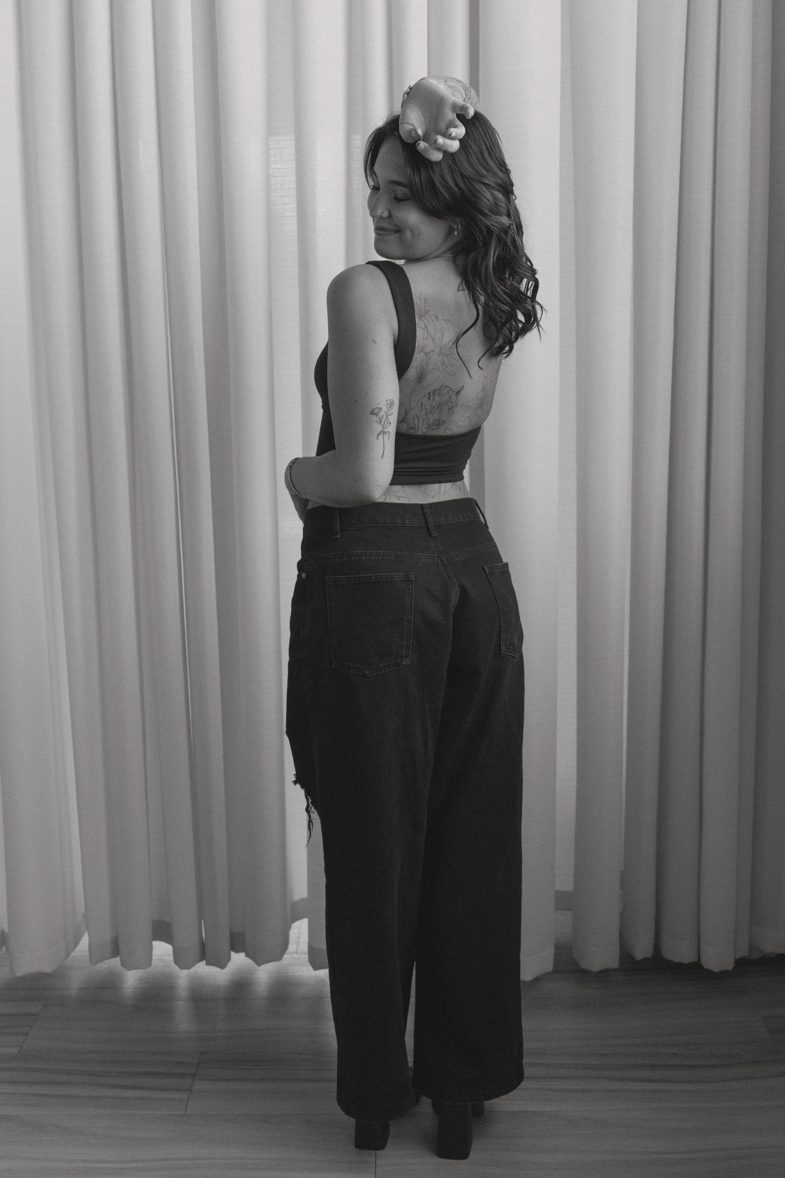 A woman with long wavy hair wearing a sleeveless top and high-waisted jeans, standing in front of curtains, with a life-sized model of a human heart on her head. She has tattoos on her arm and back and is smiling with her eyes closed.