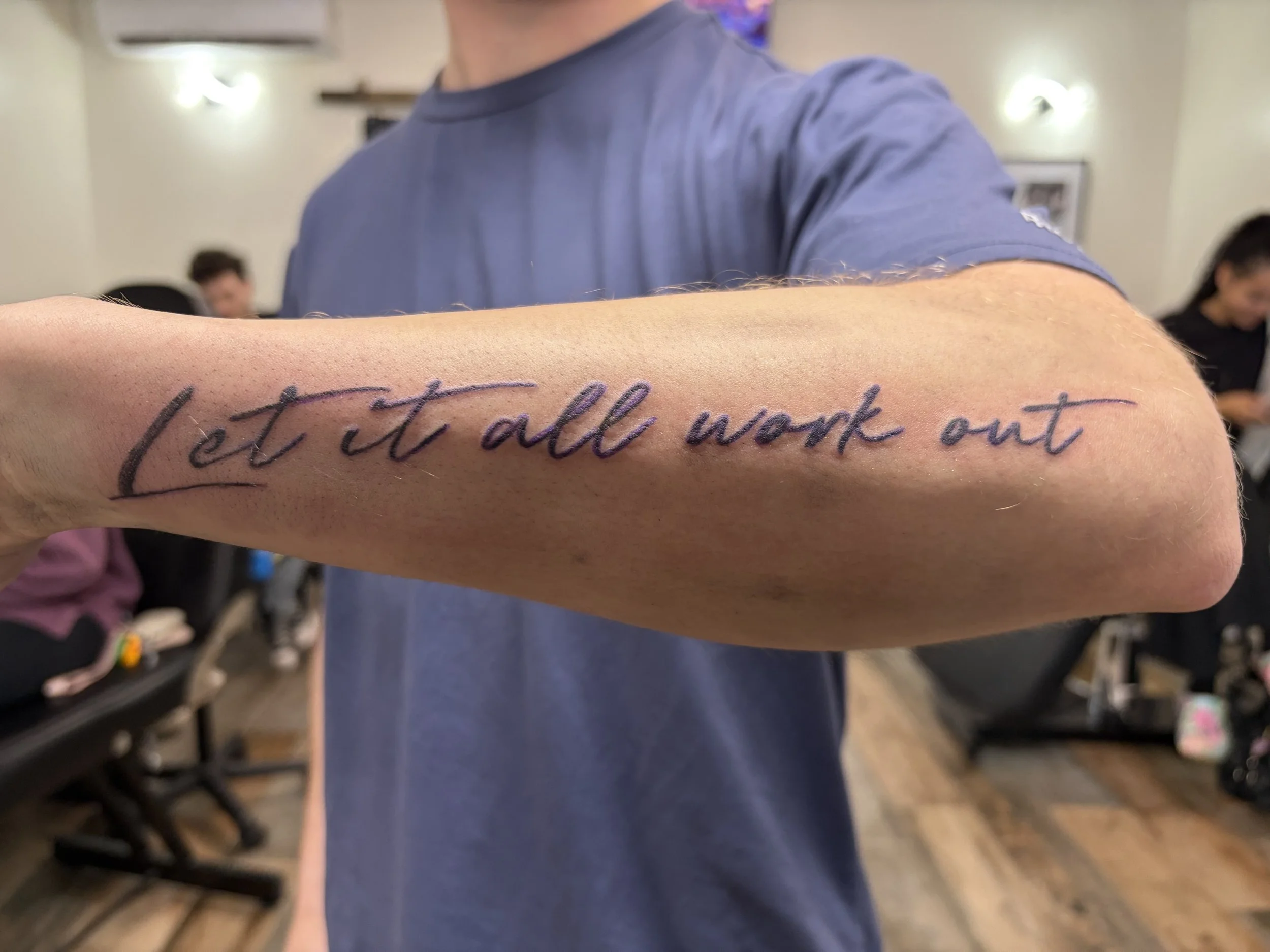 Close-up of a person's inner arm with a tattoo that reads 'Let it all work out'.